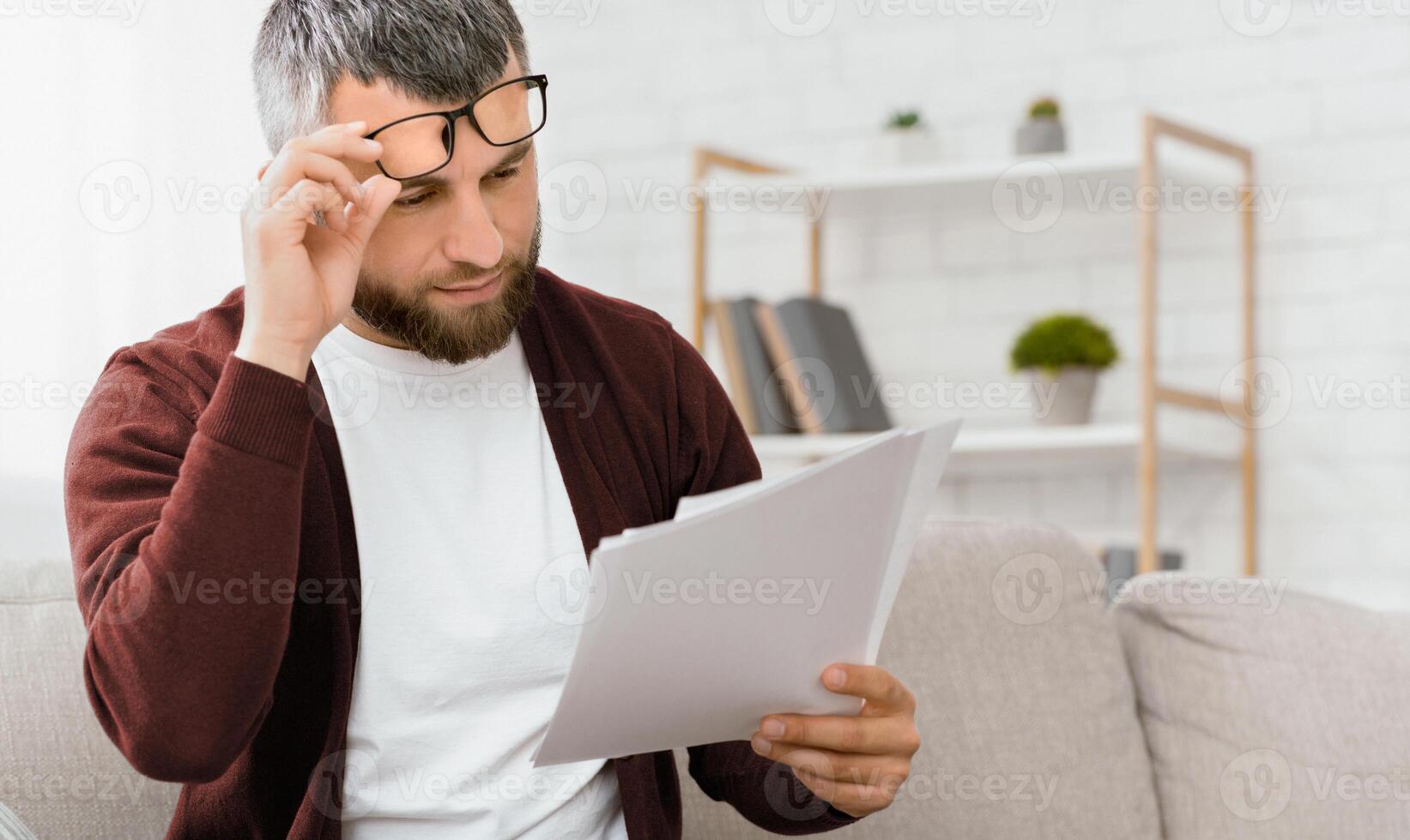 A bearded man, wearing glasses and a maroon cardigan, is carefully reading through documents while seated on a light-colored couch in a well-lit, modern living room. photo