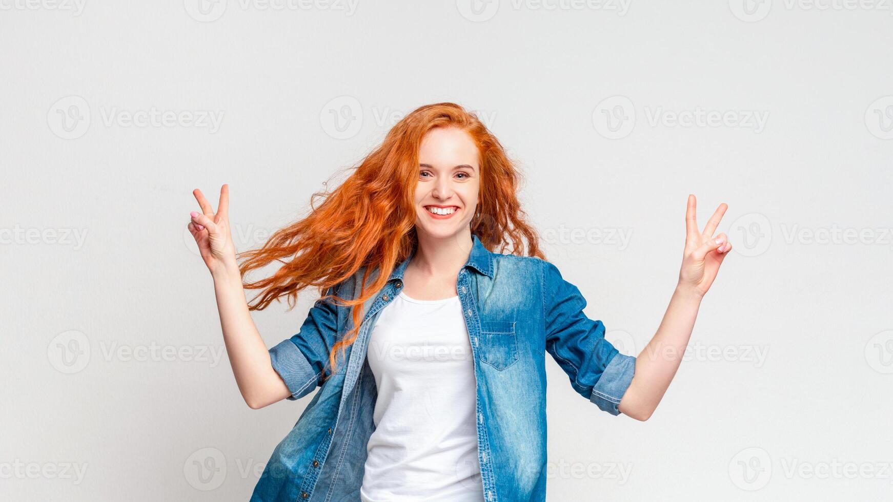 A joyful young woman with flowing red hair and a denim jacket stands against a plain white backdrop, beaming a wide smile as she playfully raises both hands to display peace signs. photo