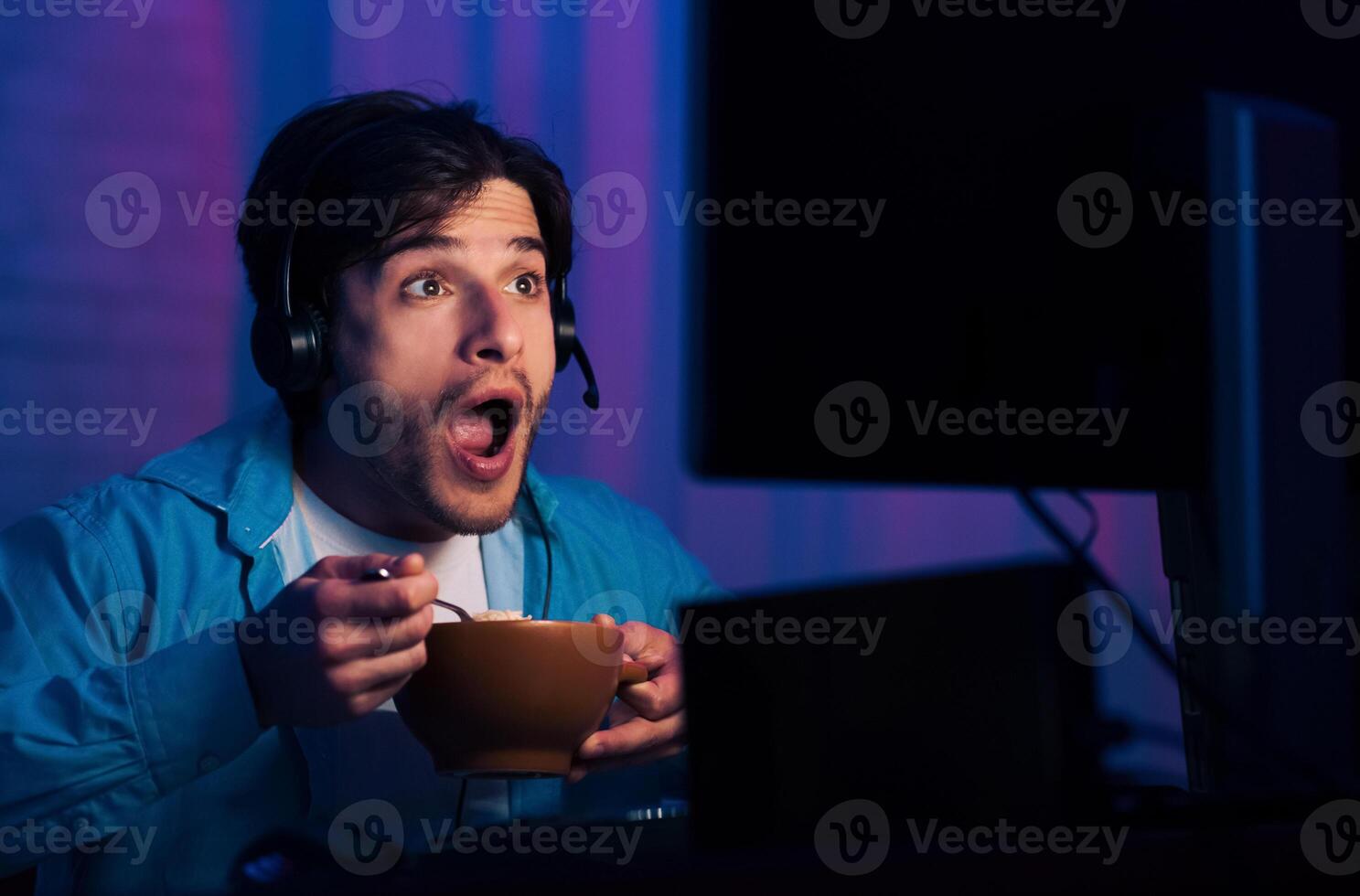 A young man wearing a headset and a blue shirt sits in front of a computer screen, looking at the screen with a shocked expression on his face while holding a bowl of cereal and a spoon in his hands photo