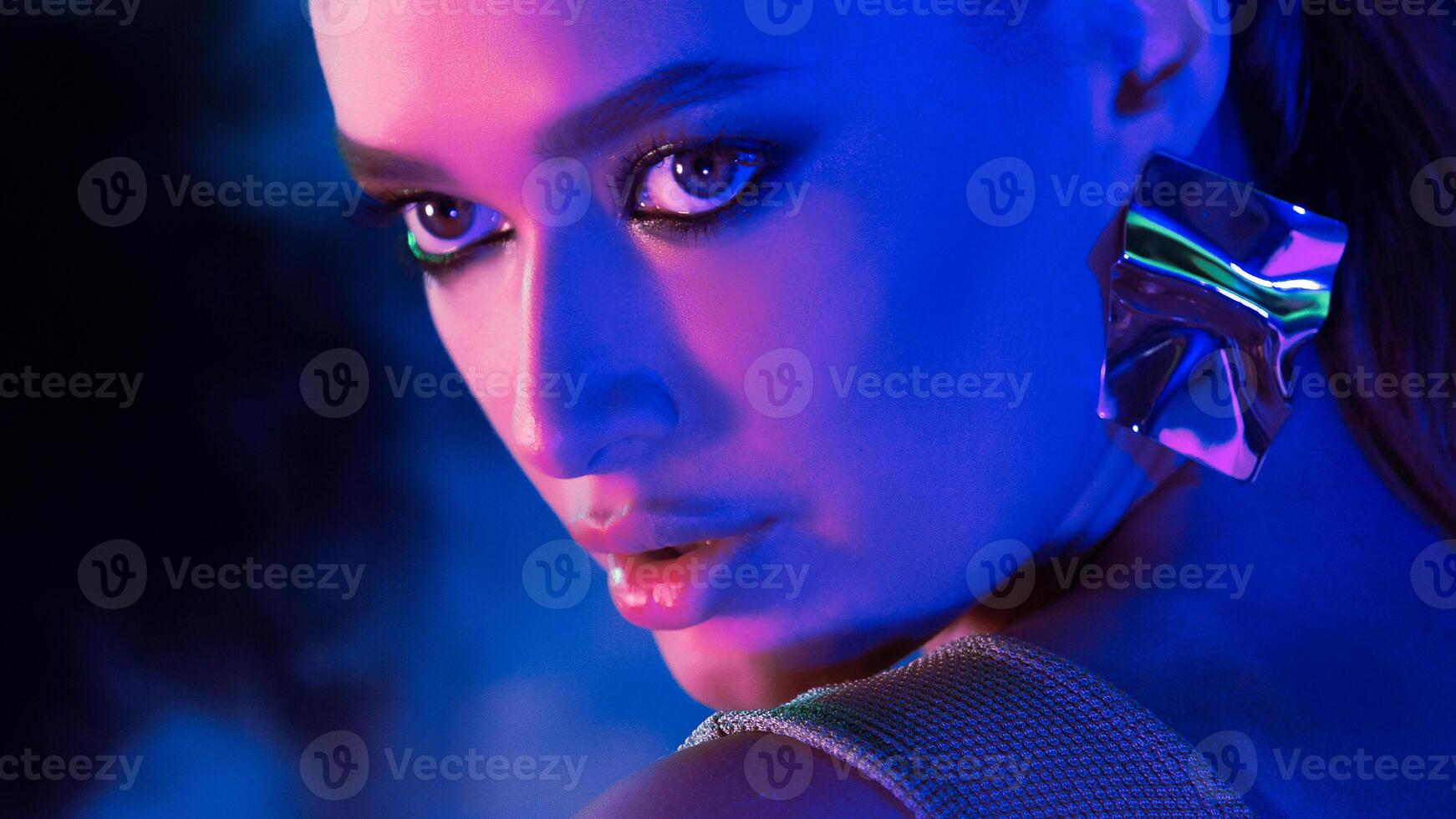 A woman stares intensely at the camera under dramatic blue and purple studio lighting. She wears bold makeup and a large metallic earring. The setting suggests a nighttime atmosphere photo