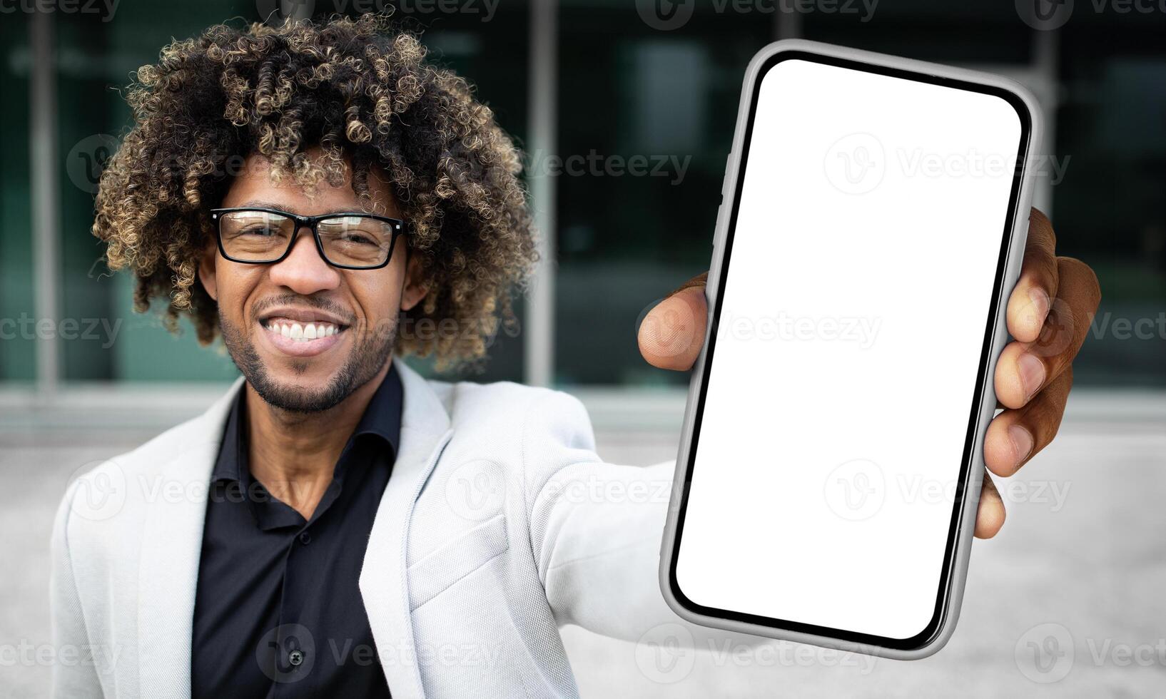 Black man standing and holding a smartphone in front of him, displaying a blank white screen. The mans hand is clearly visible as he holds the device upright. photo