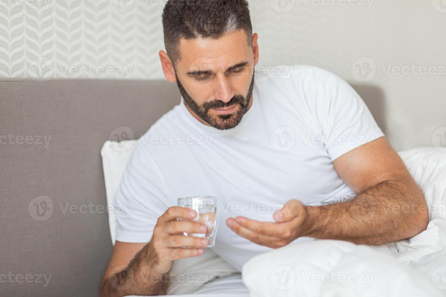 A man sits in bed, wearing a white t-shirt, holding a glass of water in one hand and a pill in the other. He appears to be about to take the pill. photo