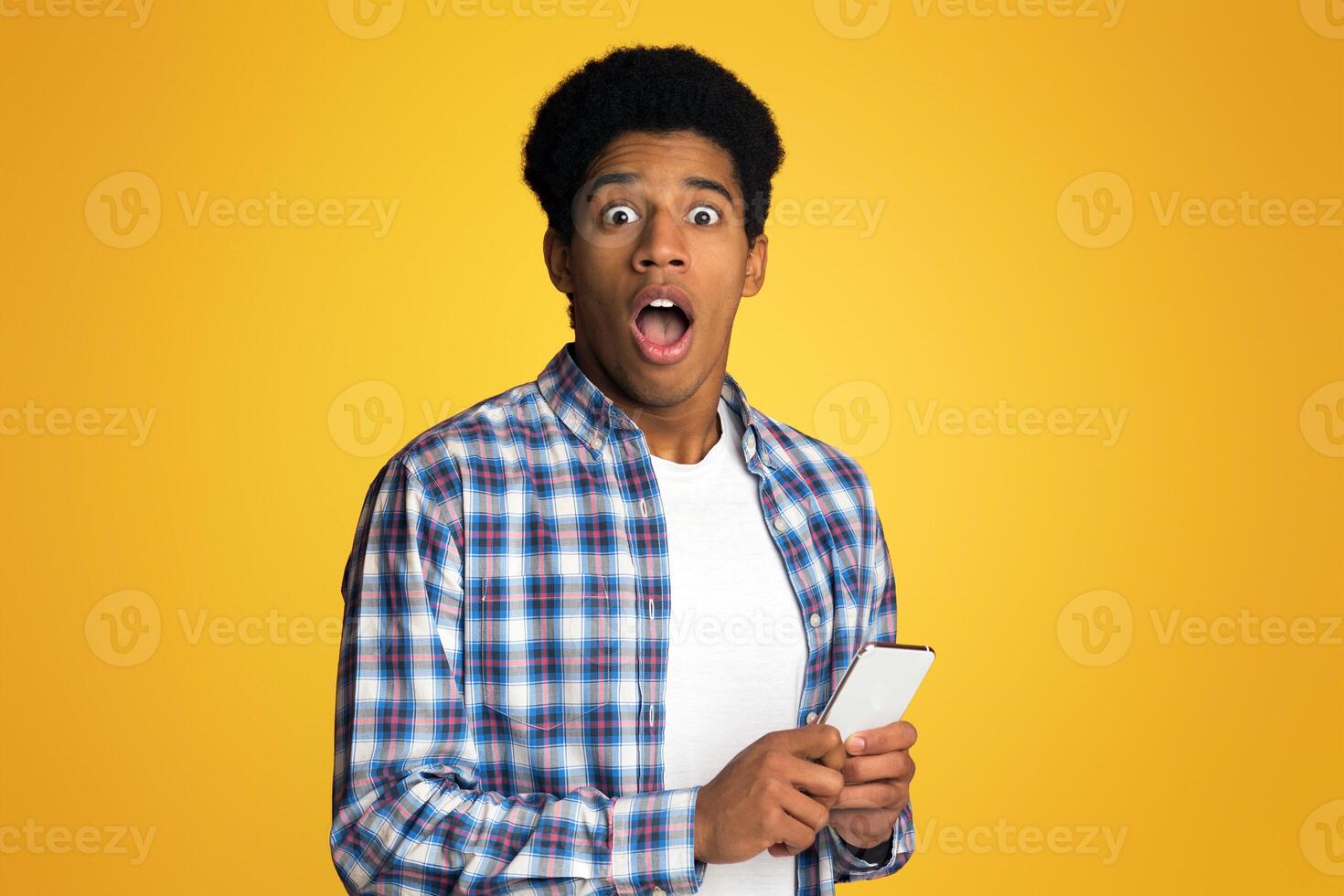 Shocked guy holding phone, astonished by social media online news, yellow studio background photo