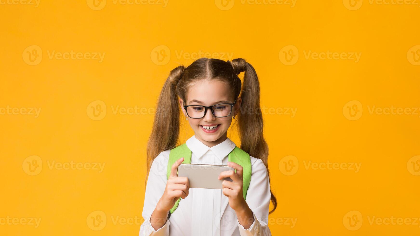 Joyful School Girl Playing Mobile Game On Phone During School Break Standing Over Yellow Studio Background. School Leisure Concept photo