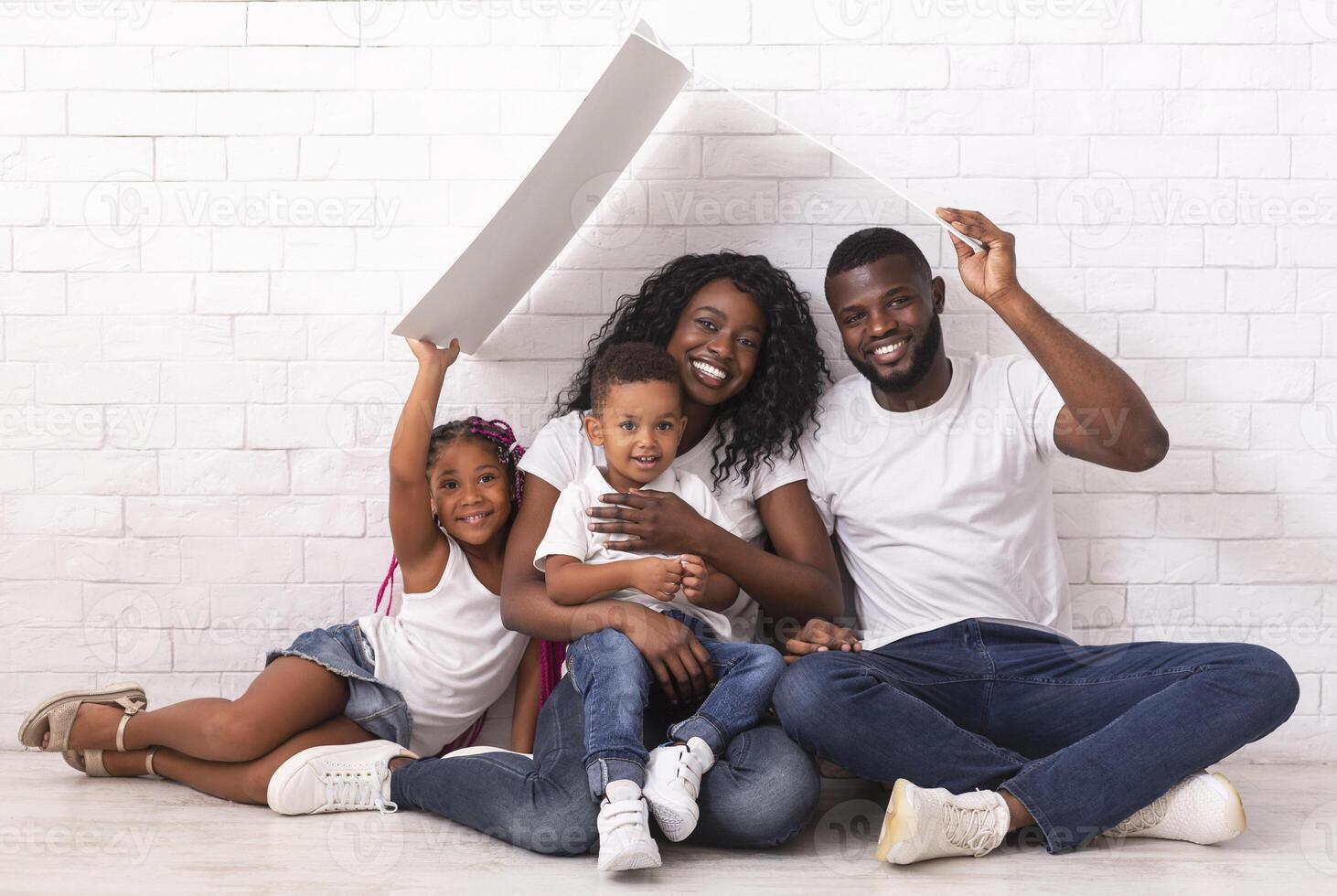 Mortgage For Young Families. Happy Black Parents And Their Two Kids Sitting Under Symbolic Roof Dreaming Of New Home Over White Wall Background photo