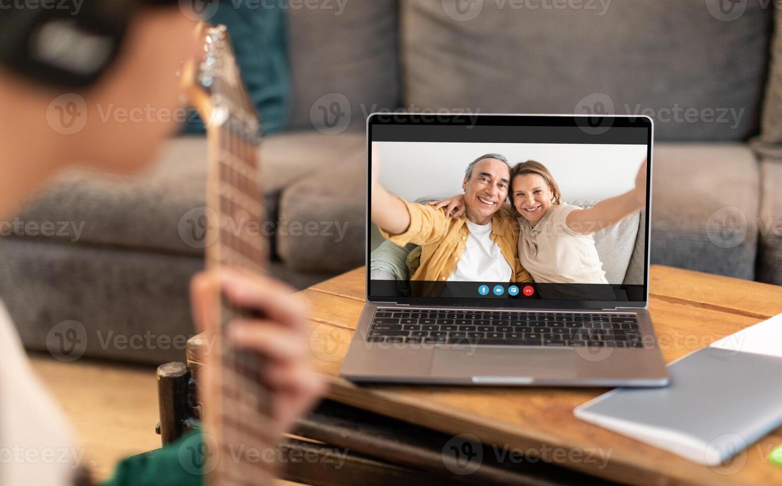 A young guy is seen playing an acoustic guitar while chatting with an older couple on a laptop. The couple is smiling and waving, seemingly enjoying the music. photo
