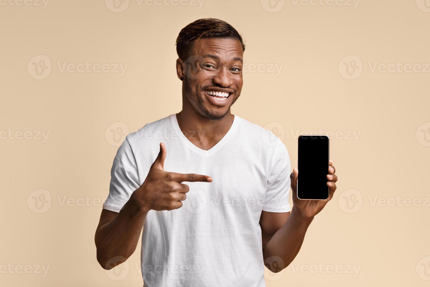 African American Man Holding Smartphone Pointing Finger At Empty Screen Standing On Yellow Studio Background. Check This Out. Mockup photo