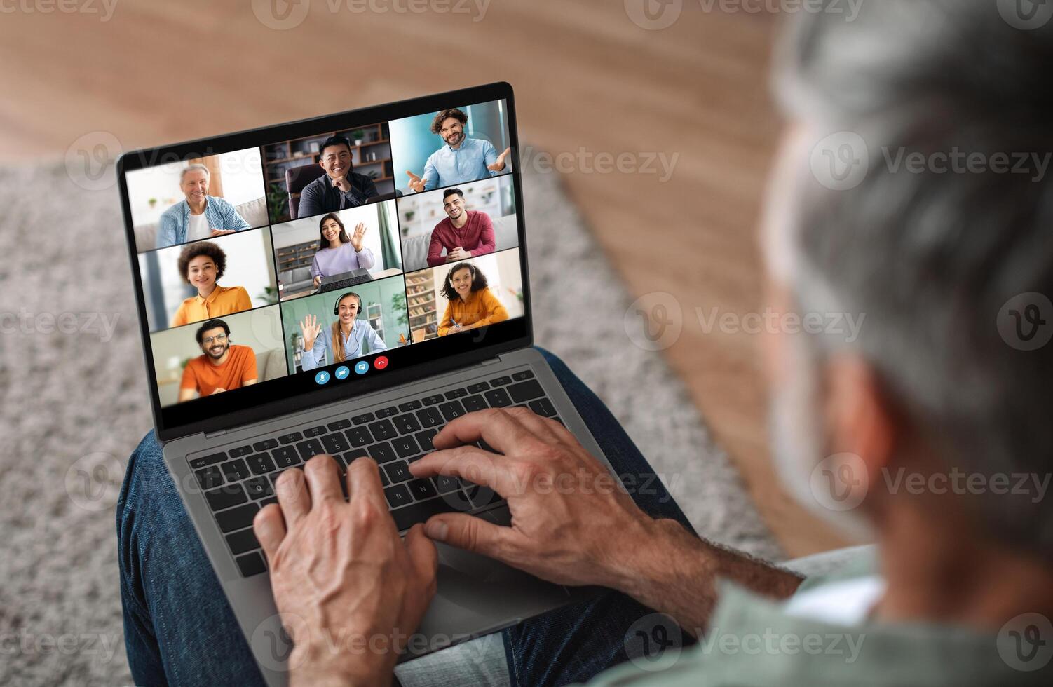 Mature man sits on a couch at home, using a laptop to participate in a virtual meeting. He is focused on the screen, which displays a grid of feeds of other participants photo
