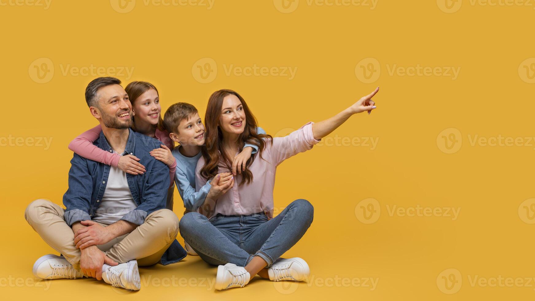 A family consisting of parents and two children is sitting on the floor indoors. They are all pointing at an object or an event happening in front of them. photo