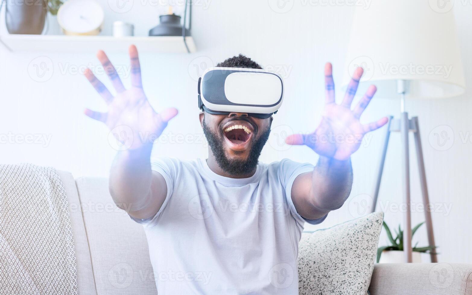African American man wearing a virtual reality headset and interacting with the virtual world. He is sitting on a couch and has his hands extended outwards as if he is reaching for something photo