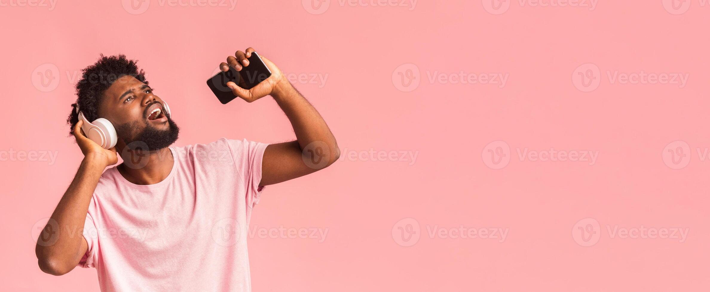 Funny afro guy with wireless headset singing over pink background, using karaoke mobile app photo