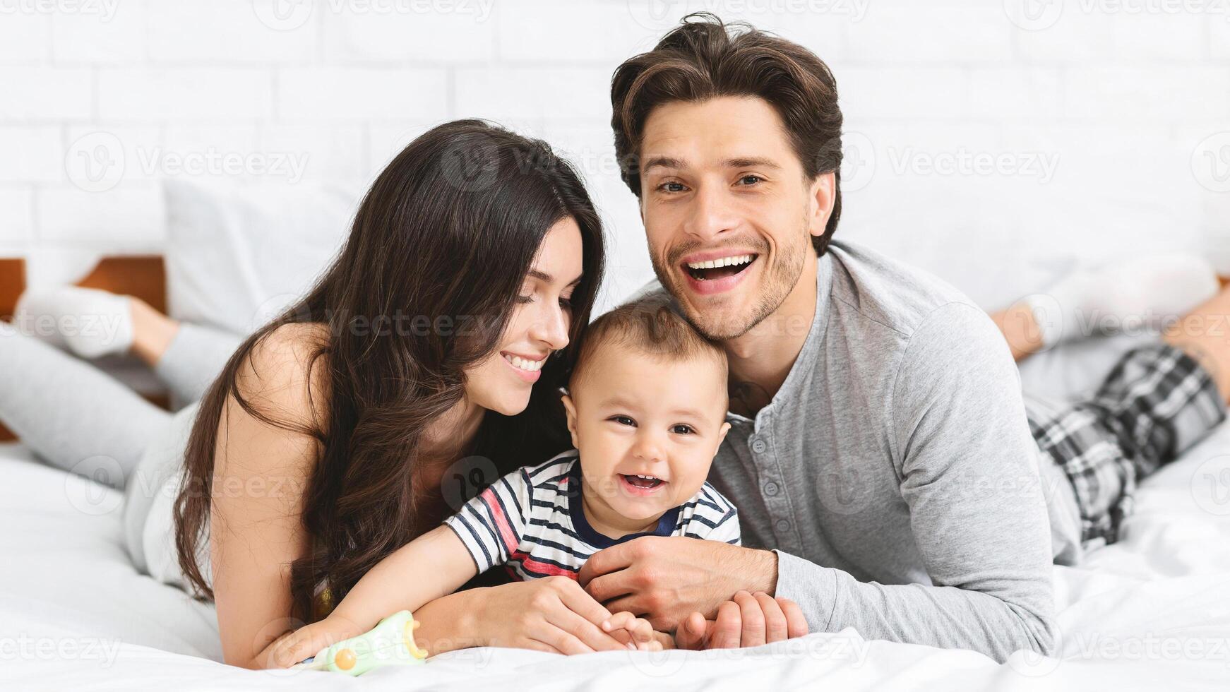 This image shows a happy family of three, a man, woman, and baby, lying on a white bed. The parents are smiling, and the baby is laughing. They are all wearing casual clothing photo