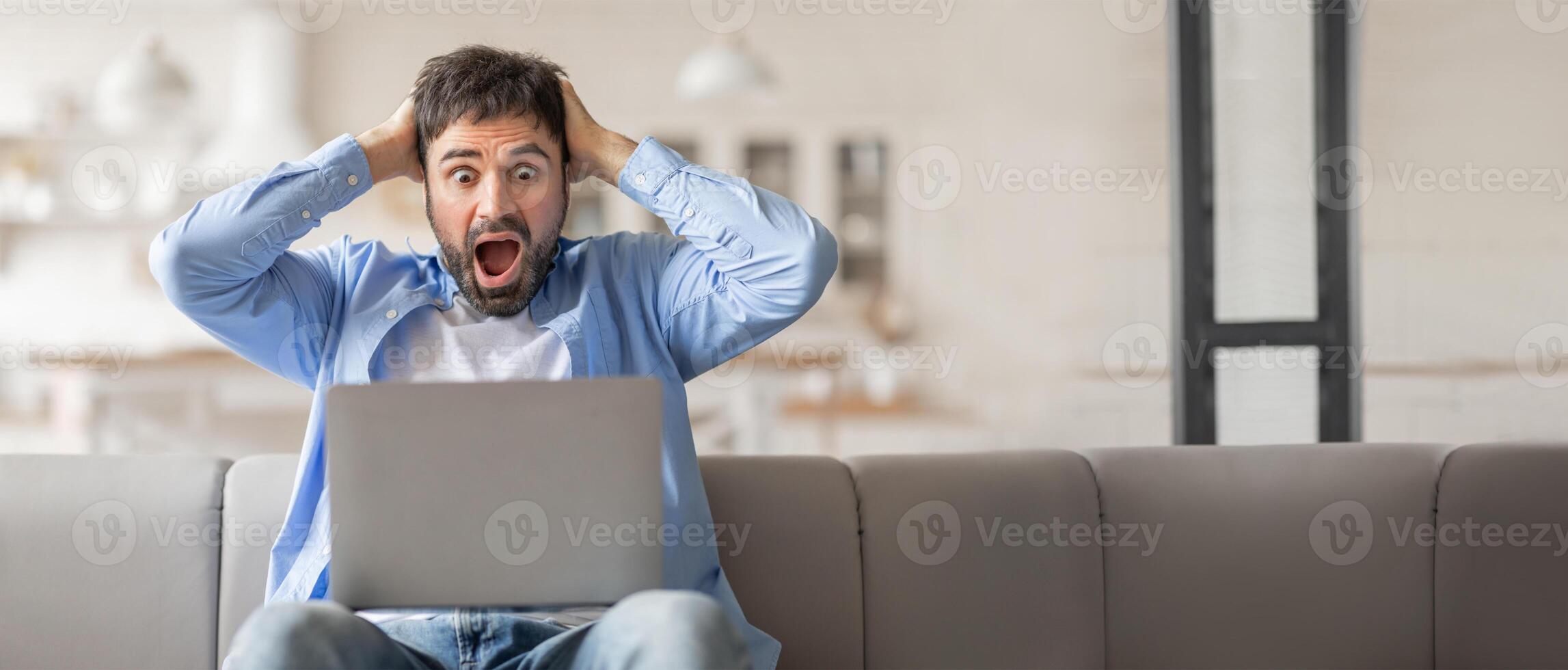 A man sitting on a couch at home is holding his head in frustration while staring at his laptop computer. He shows a mix of shock and stress during a challenging work moment. photo