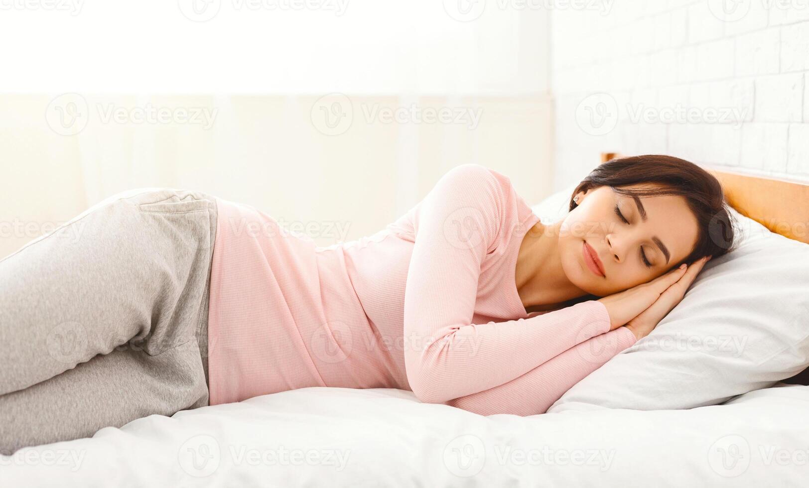 A woman is lying down on a bed, resting her head on a soft pillow. She appears comfortable and relaxed in the cozy bedroom setting. photo