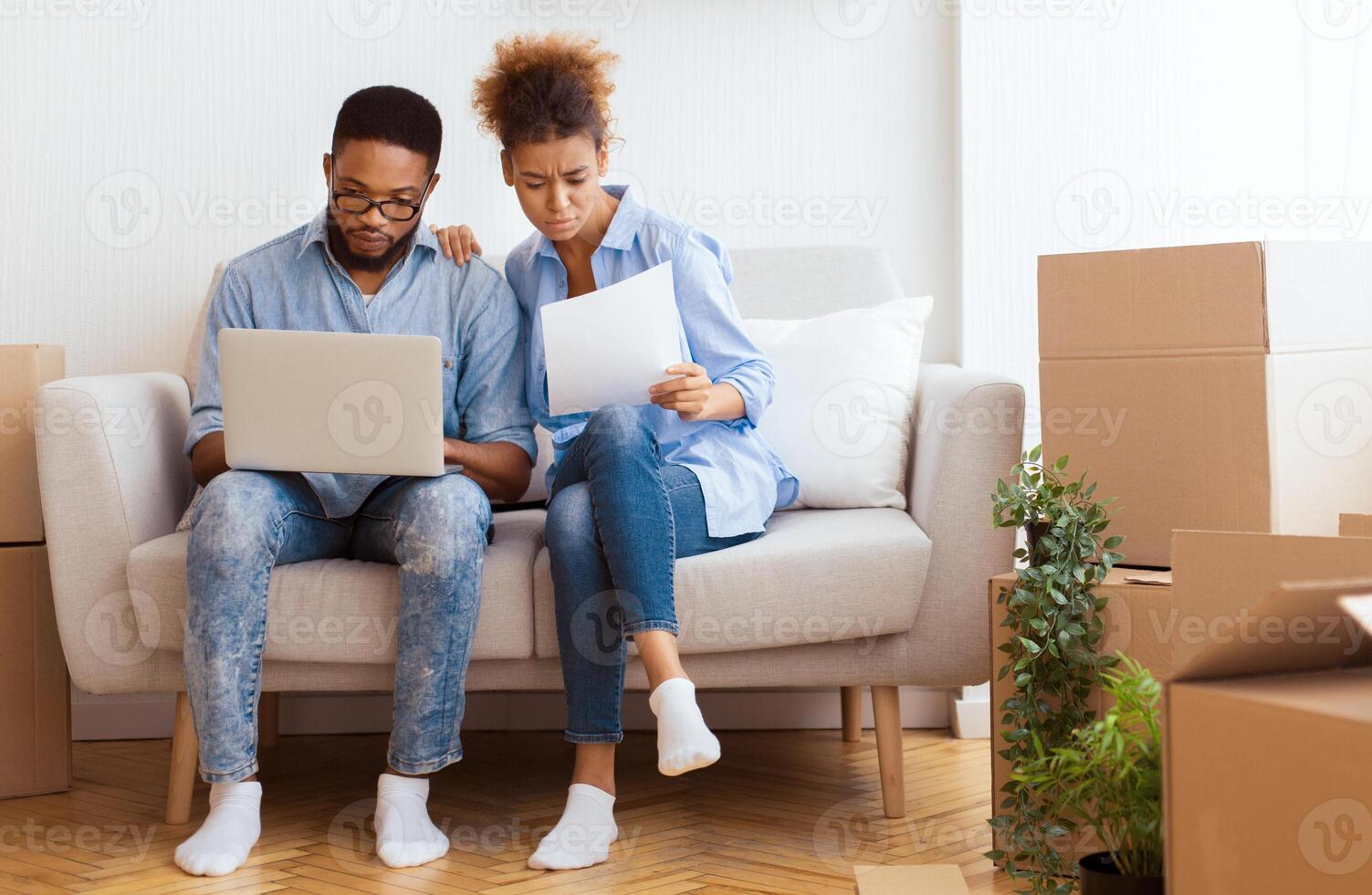 Buying House. Serious Black Couple Using Laptop Reading Property Ownership Papers Sitting On Sofa In New Home. Empty Space photo