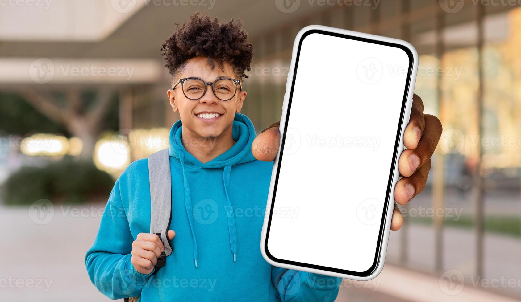 A young black guy with curly hair and glasses is smiling at the camera while holding up a smartphone with a blank screen, standing in front of a university building photo