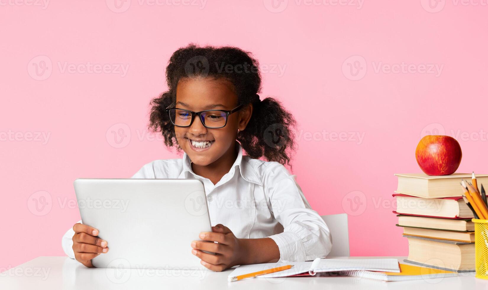 Smiling Afro Schoolgirl Using Tablet Computer Sitting At Desk Over Yellow Studio Background. School And Gadgets Concept. photo
