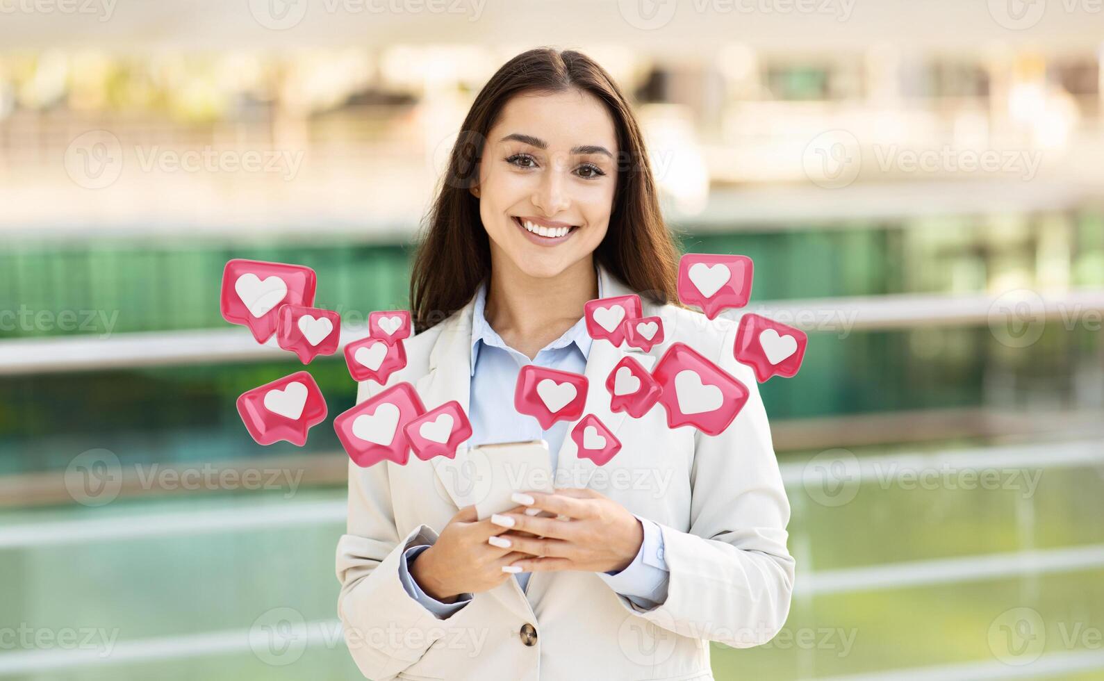 A smiling woman in a white blazer stands in a modern urban setting, holding a smartphone while surrounded by animated hearts, representing likes and notifications. photo