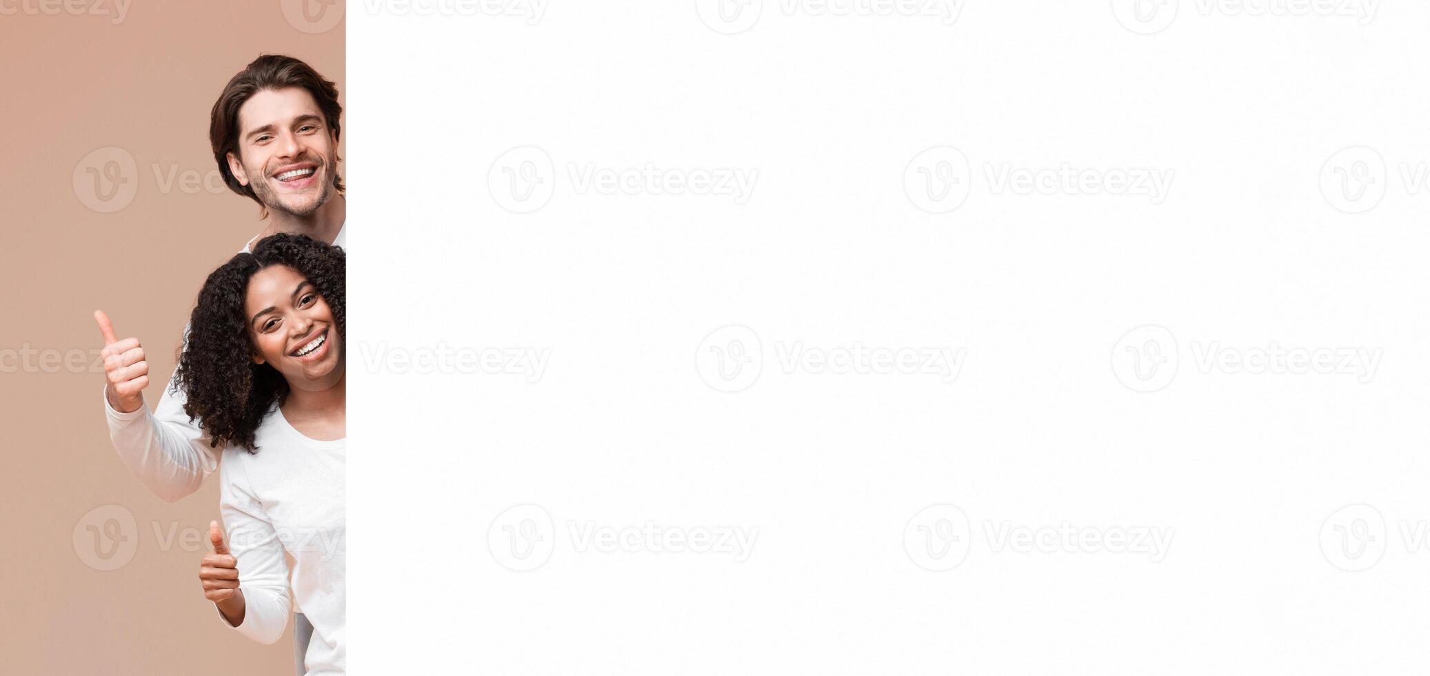 Happy multiracial couple peeking behind white advertisement board and showing thumbs up, recommending something photo
