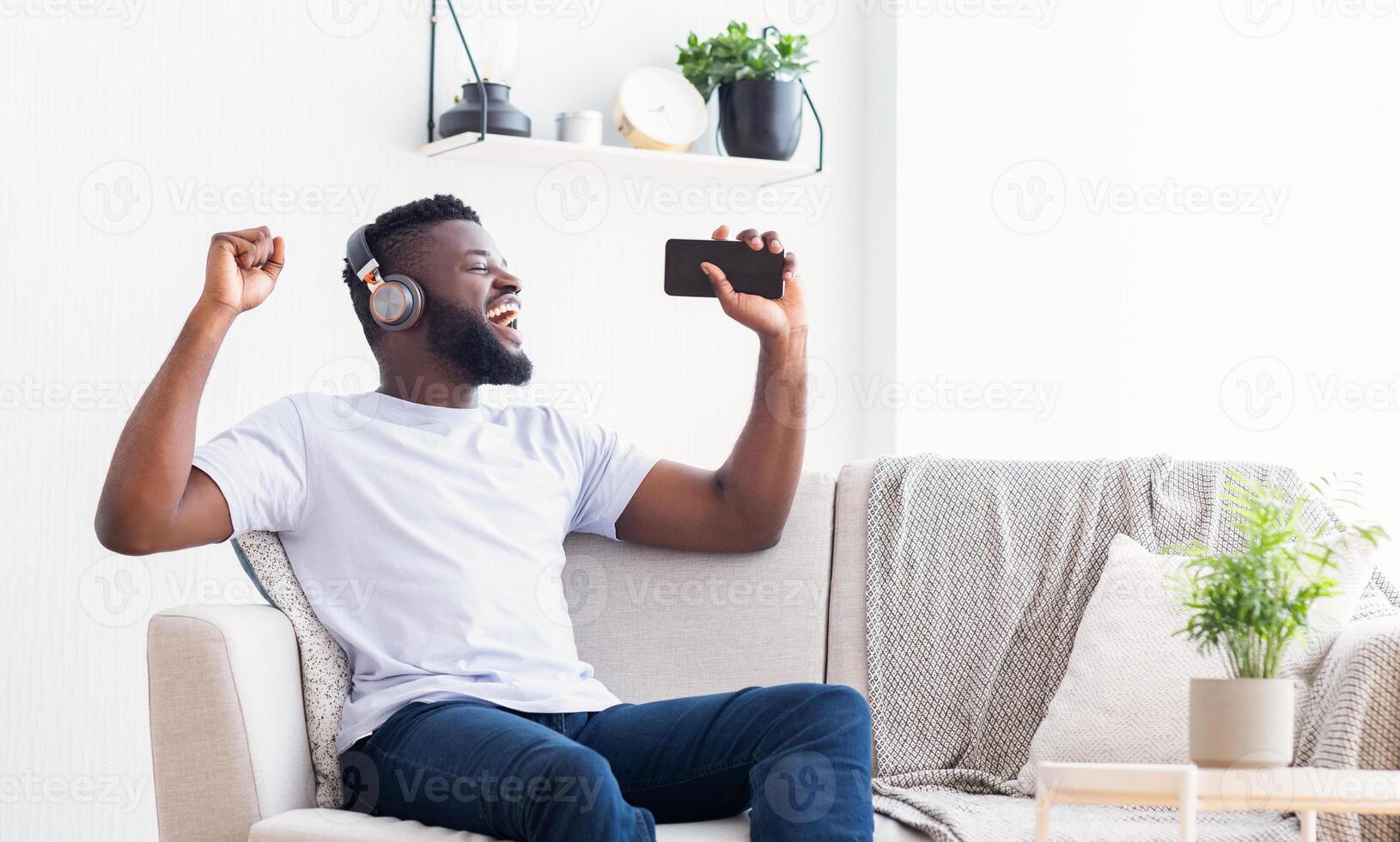 A young Black man is sitting on a white couch in his living room, wearing headphones and holding a smartphone. He is singing along to music, his face lit up with joy, copy space photo