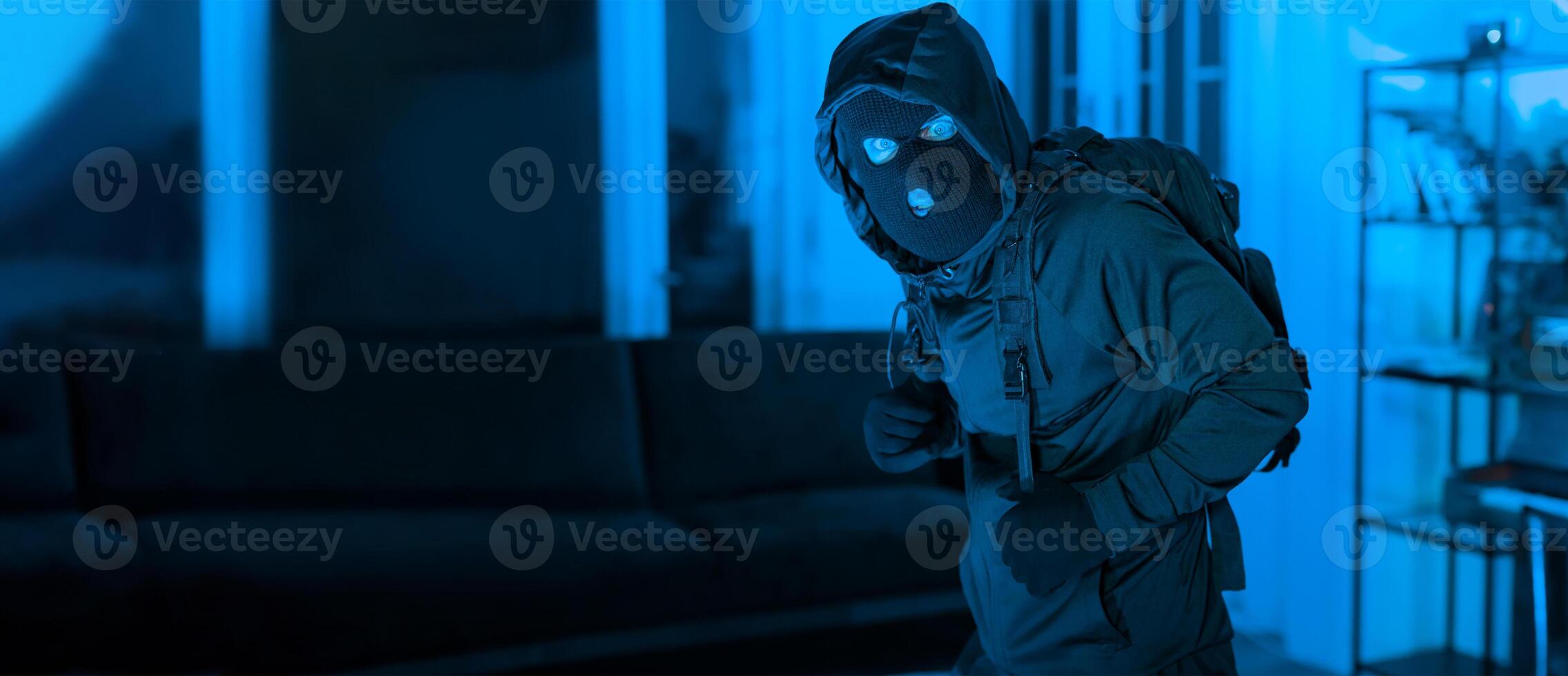 A figure dressed in dark clothing with a hood and mask stands in a dimly lit room at night. The atmosphere is tense, with blue lighting highlighting the scene. photo