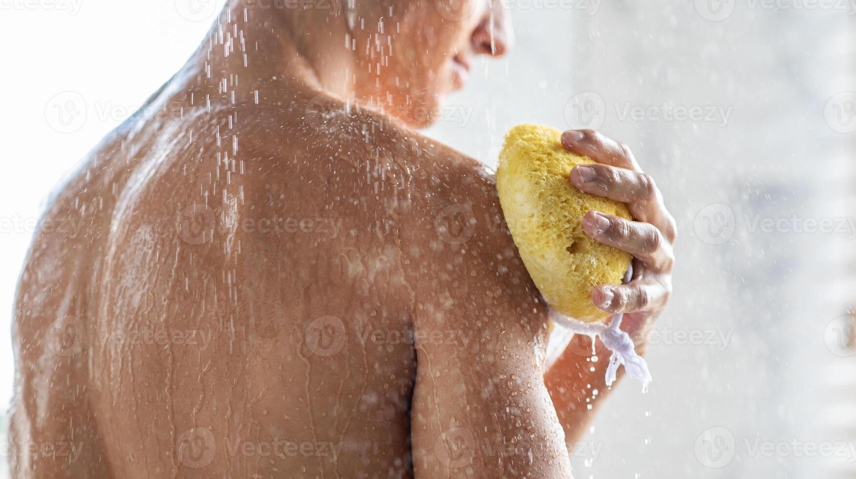 Bodycare Concept. Unrecognizable muscular man taking refreshing shower and washing his hands and shoulders with sponge, back view. Guy standing in the bathroom under falling hot water drops photo