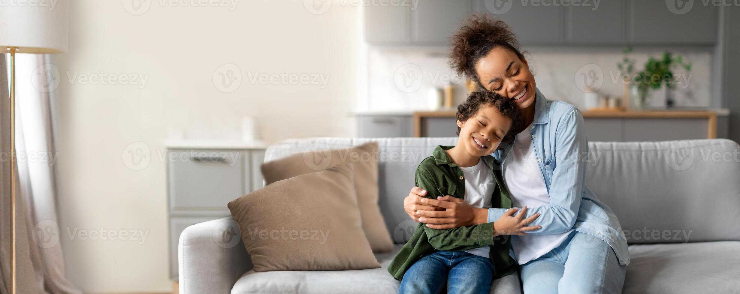 Black mother and her son sit together on a comfortable sofa, smiling and hugging each other. The living room features soft colors and a warm atmosphere, creating a sense of love and happiness. photo