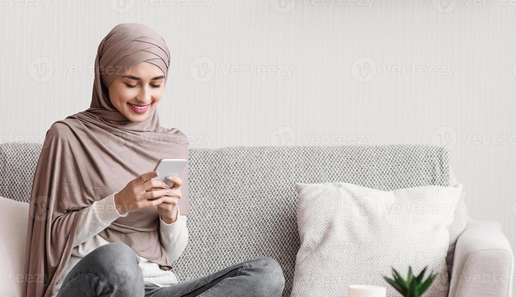 Smiling muslim girl in headscarf using smartphone at home, messaging or browsing social networks, relaxing on couch in living room, copy space photo