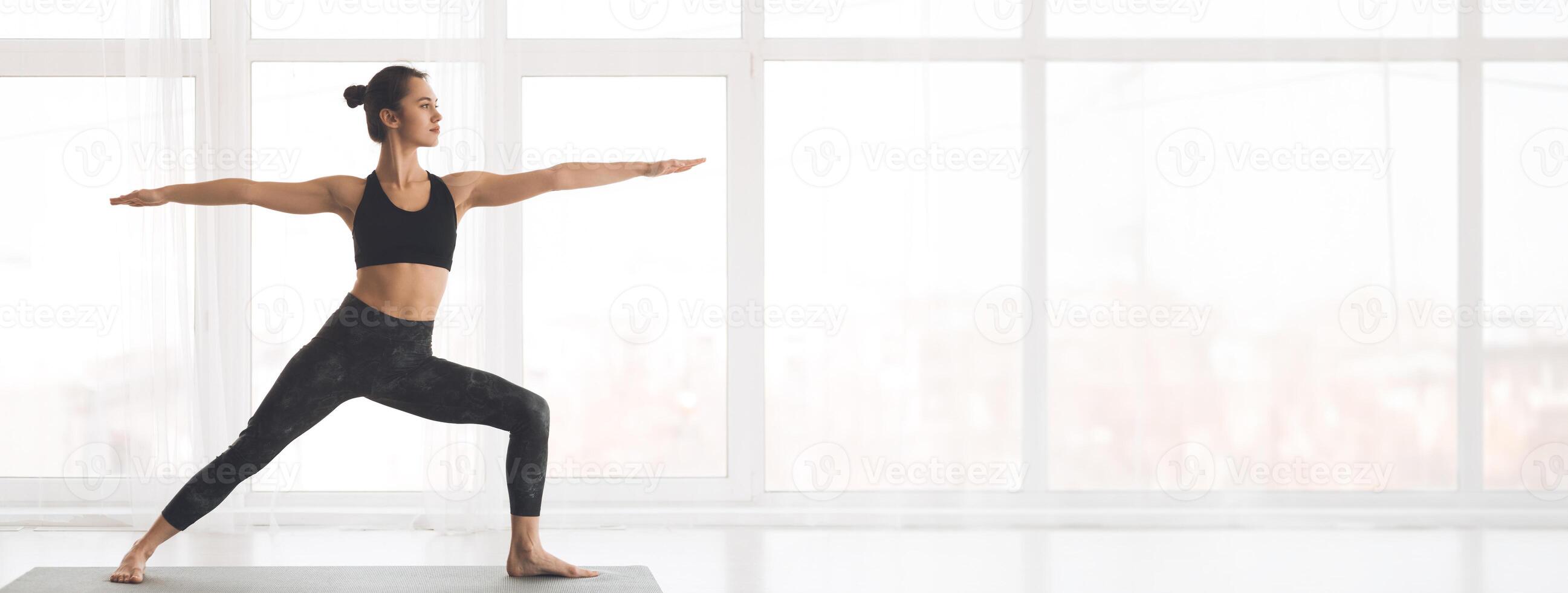 A person performs the warrior pose in a well-lit studio, showcasing strength and balance against a backdrop of large windows, copy space photo