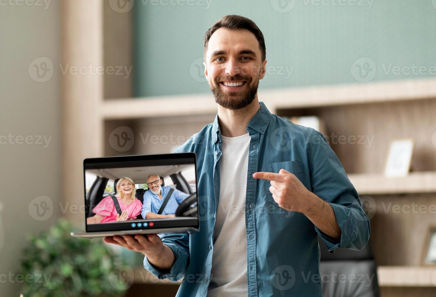 A man stands in a home office, smiling and pointing at his laptop screen, which shows a call with an elderly couple sitting in a car. photo