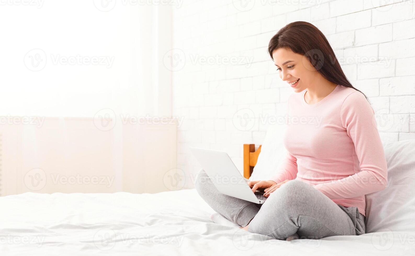 A woman is sitting on a bed, focusing on a laptop computer screen. She appears engaged in her work or leisure activity, with her hands typing on the keyboard, copy space photo