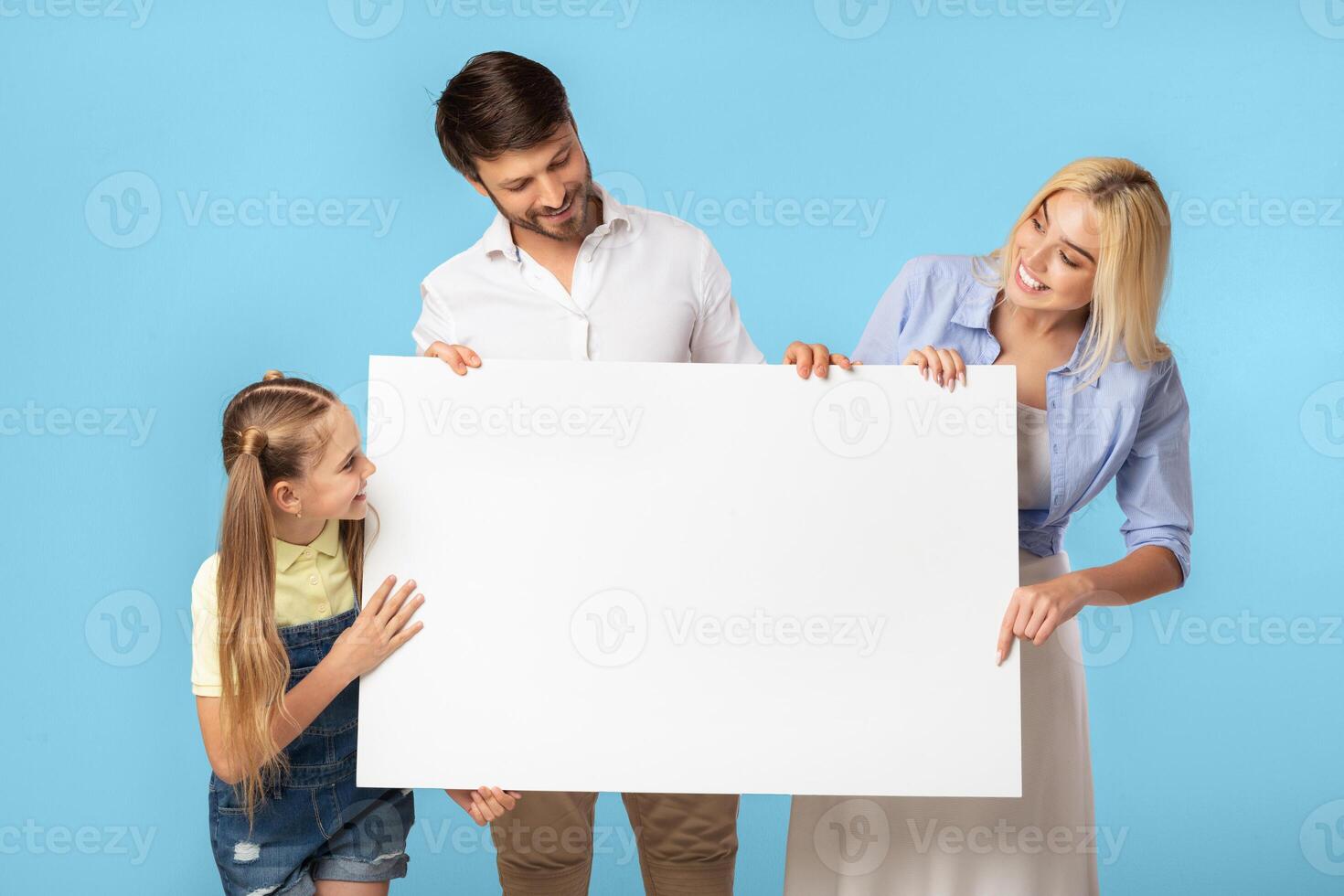 A family of three, a father, a mother, and their daughter, are standing in front of a light blue background holding a large blank white sign. photo