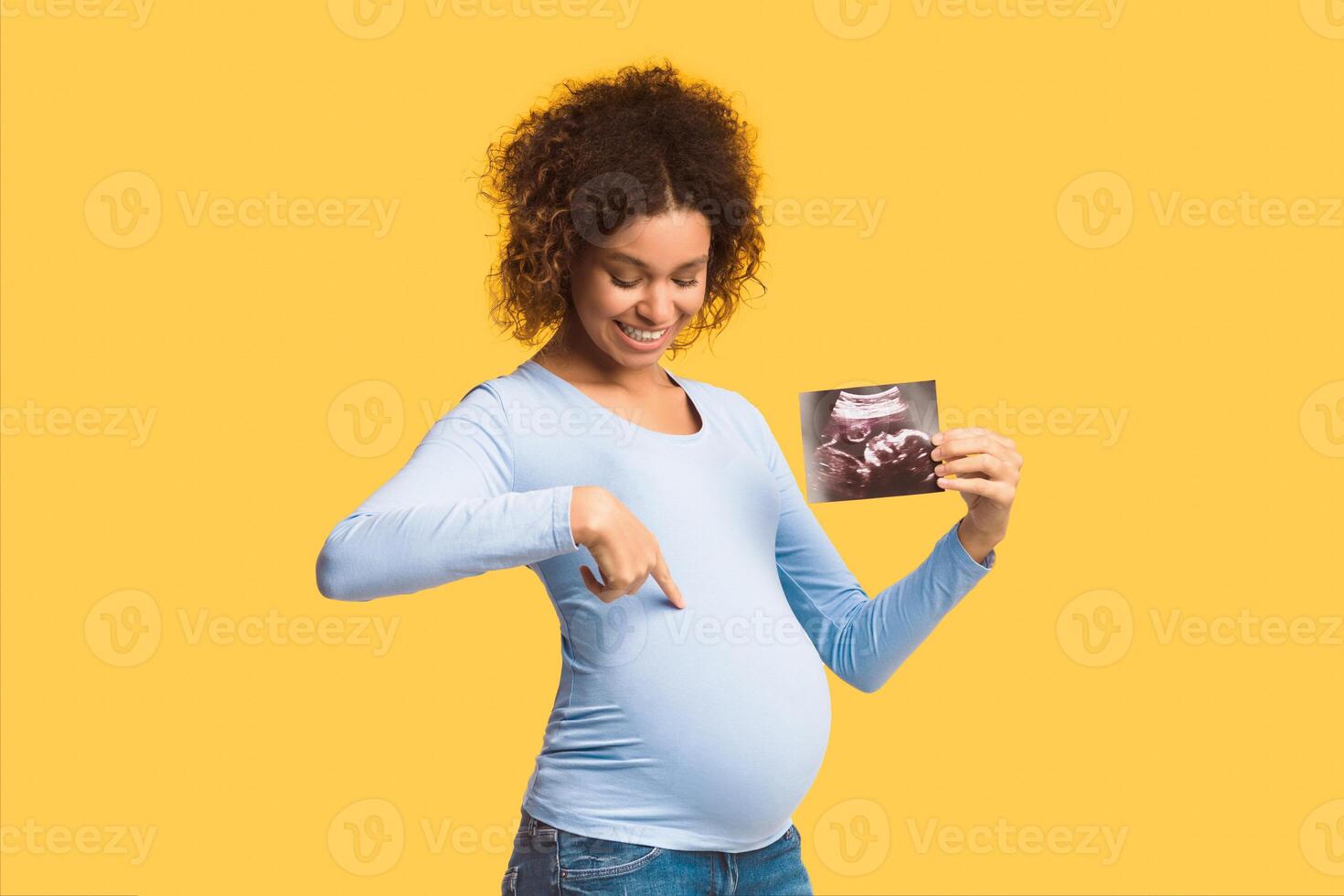 Happy pregnant lady showing ultrasound scans and pointing at belly over white background photo