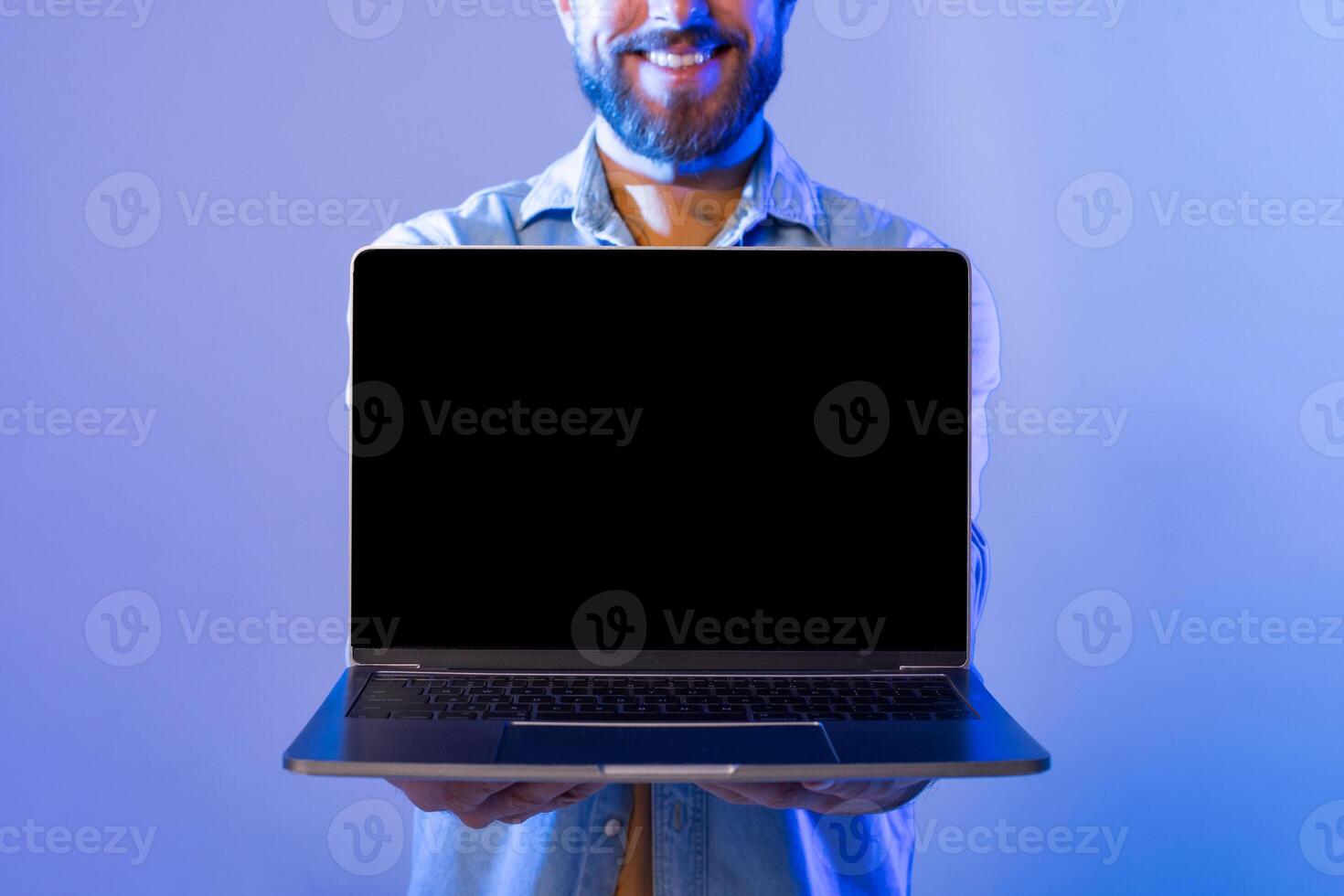 A smiling man with a beard stands against a blue background, holding a laptop with the blank screen facing forward. He appears cheerful and enthusiastic, cropped photo
