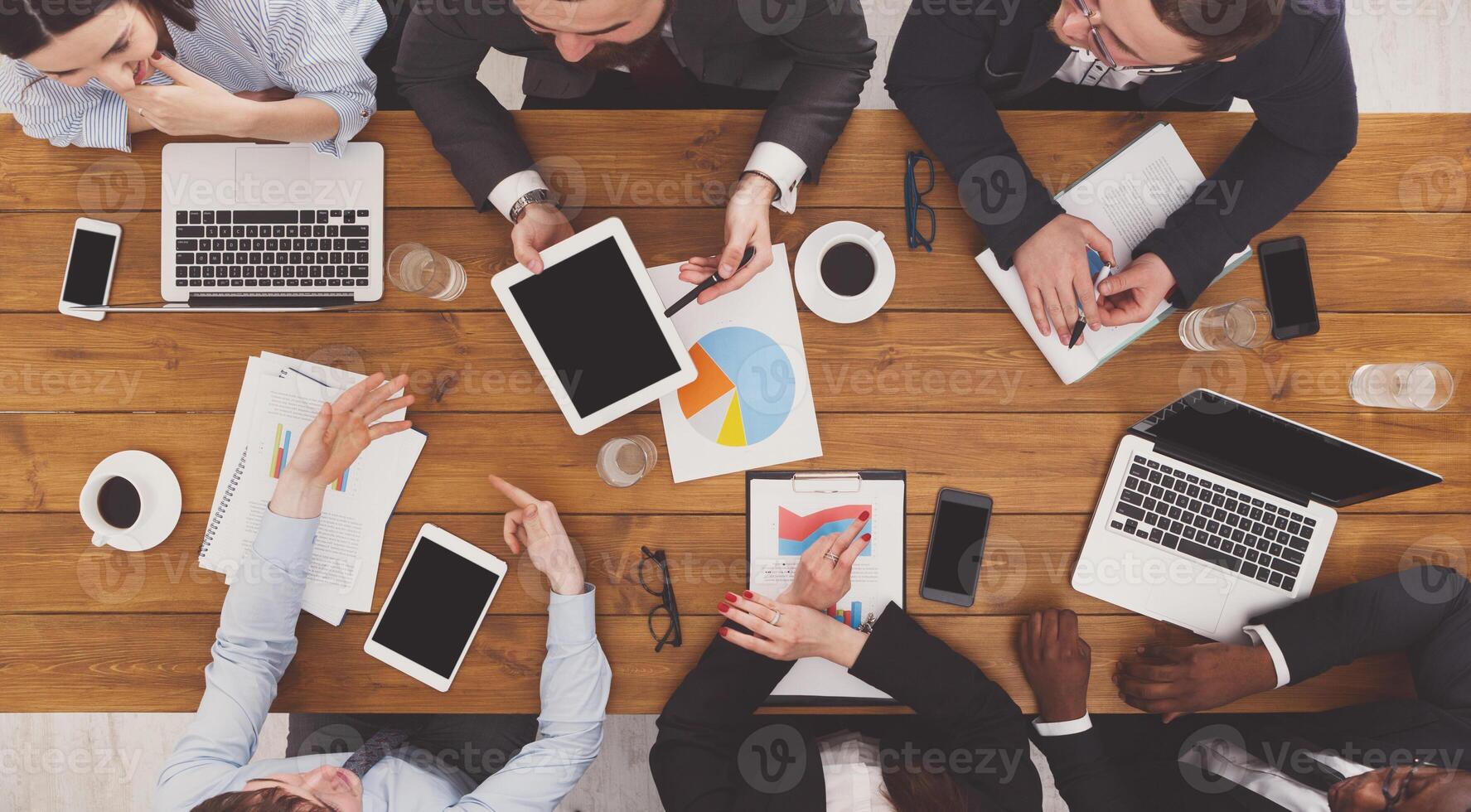 Group of busy business people working in office, top view of wooden table with mobile phones, laptop, tablet and documents papers with diagram. Men and women team together have brainstorm discussion. photo