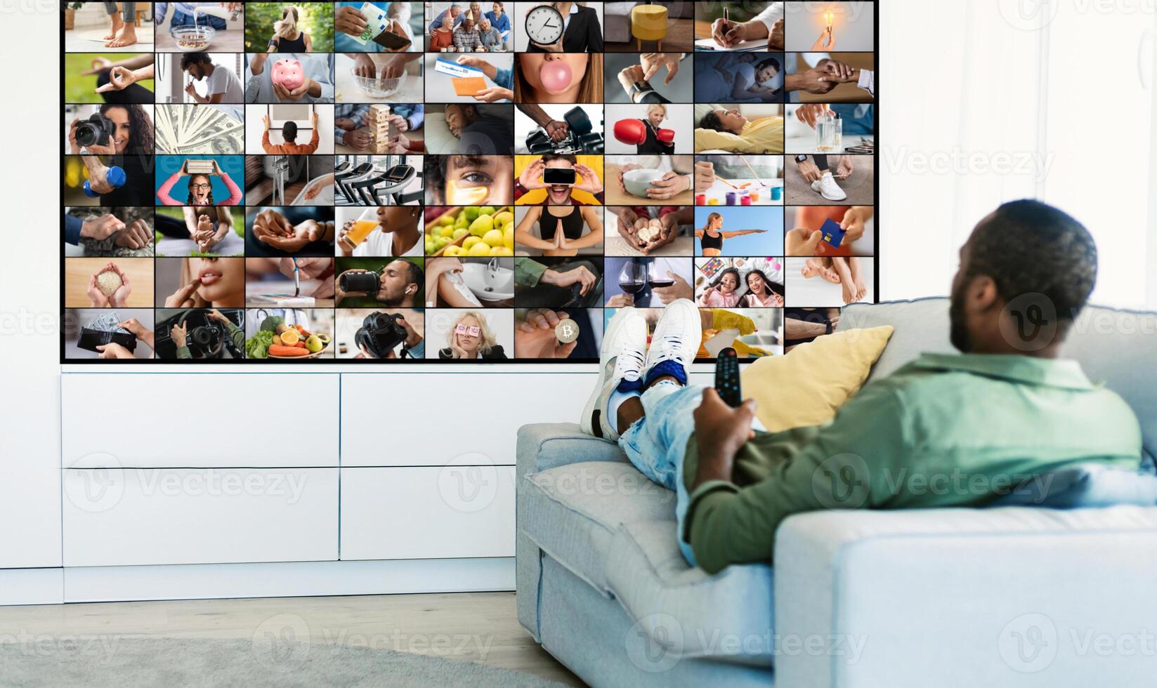 African American man is sitting on a white couch in a living room, watching television. The TV screen is filled with a diverse mosaic of smaller images, showcasing various activities and people photo