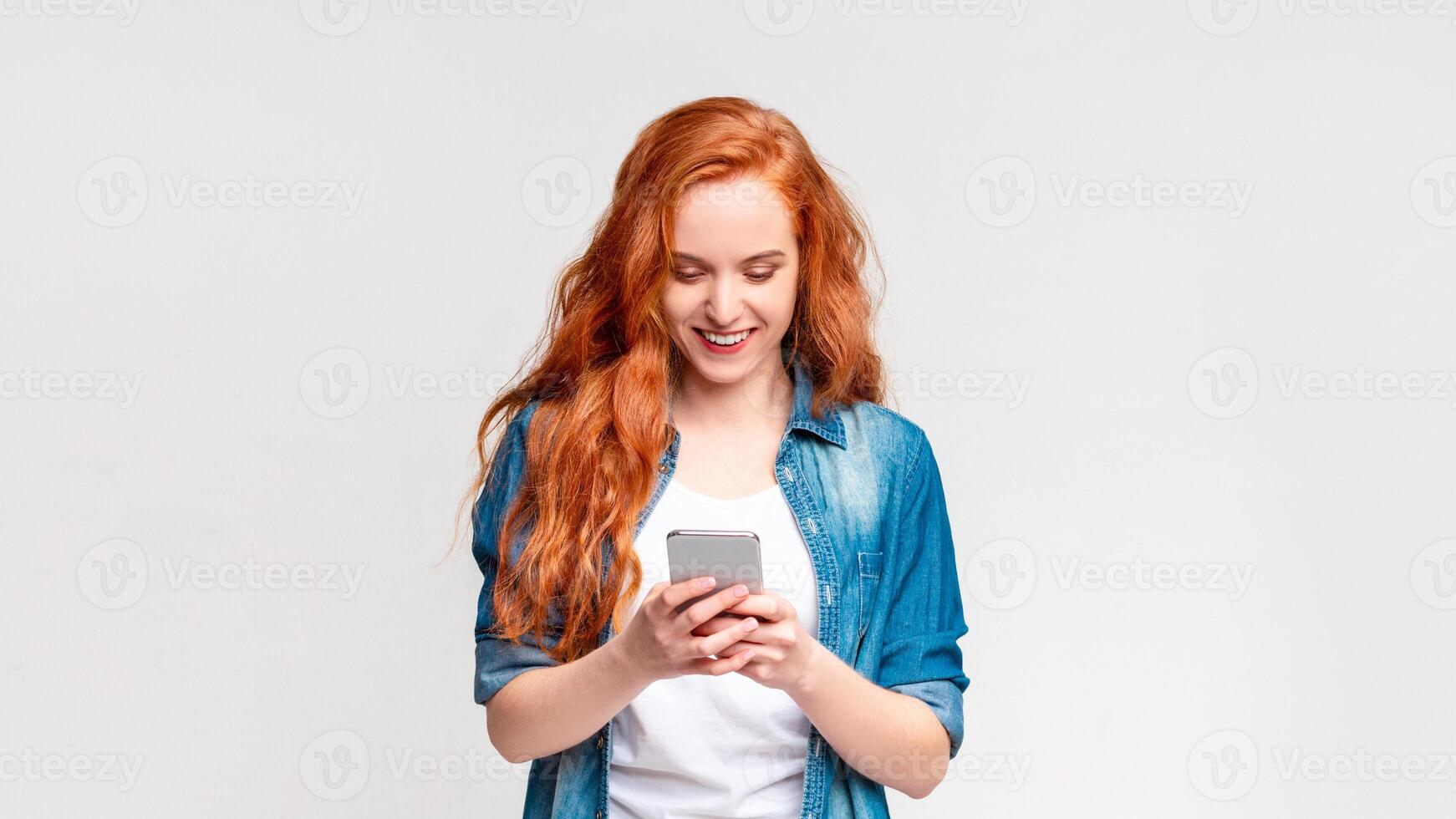 A woman with vibrant red hair is intensely focused on her cell phone screen. She appears engrossed in whatever she is viewing, holding the device with both hands photo