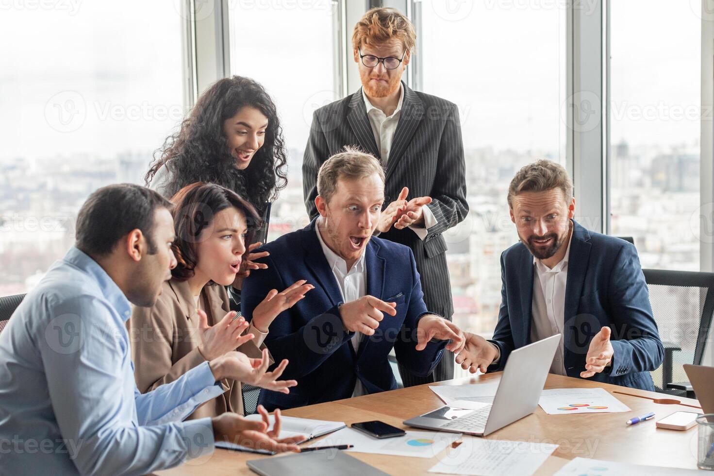 A group of business professionals gather around a laptop in a modern office setting, displaying excited reactions to the information on the screen. They are gathered around a large wooden table photo