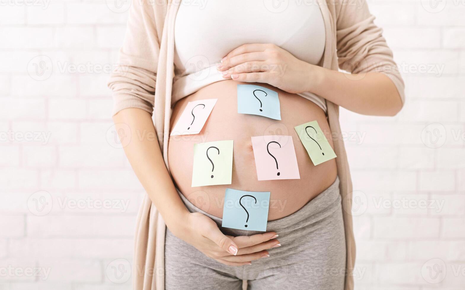 Choosing baby name. Confused pregnant woman with question marks on paper stickers on tummy photo