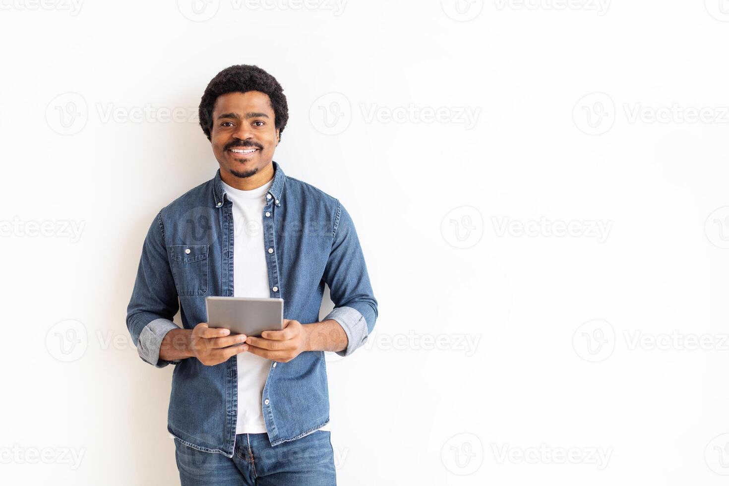 African American man is standing while holding a tablet in his hands. He appears focused on the screen, possibly reading or watching something. The background is white with copy space photo