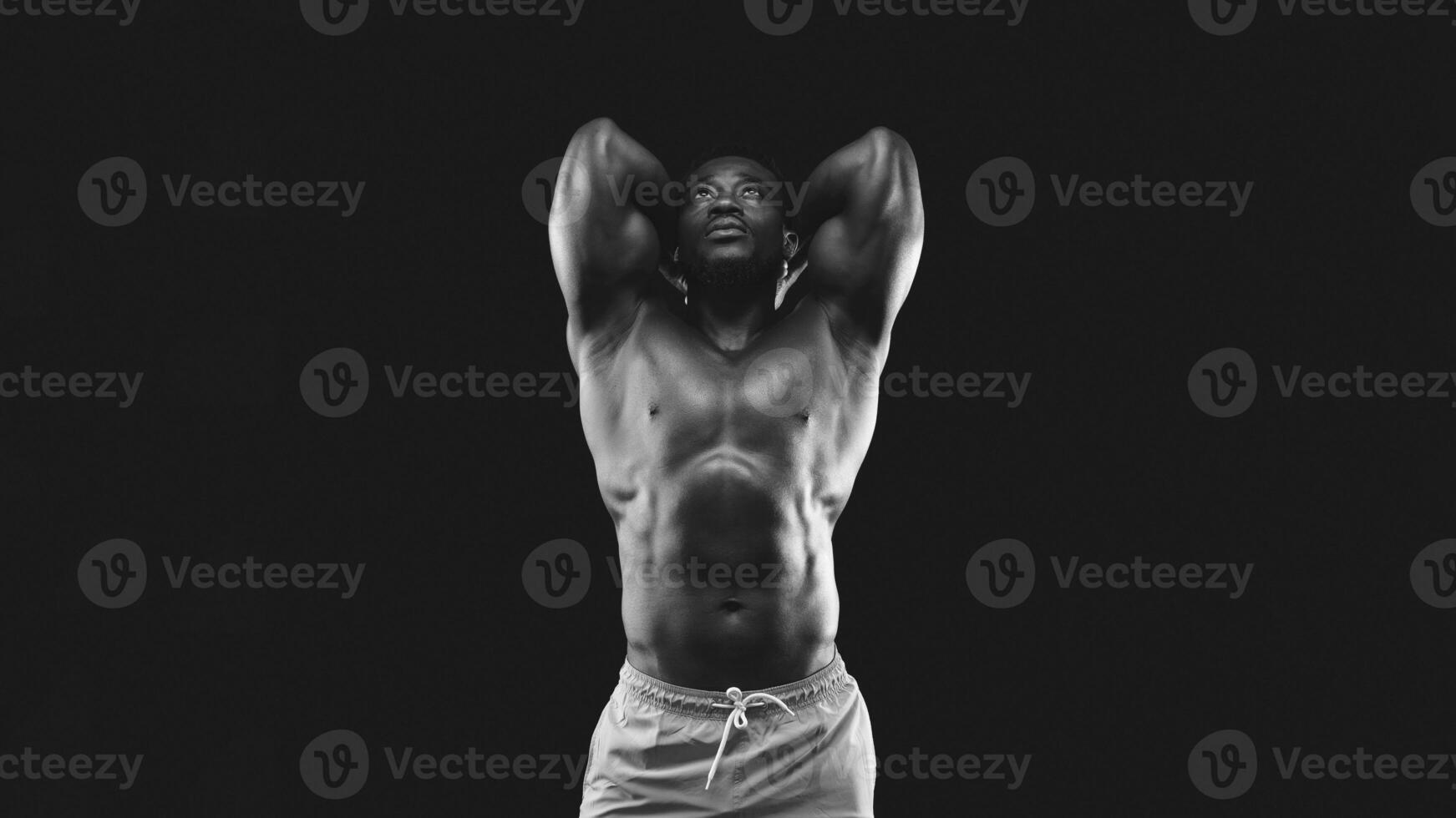 A muscular man poses with his arms raised above his head, showcasing his physique. The image is shot in black and white against a dark backdrop. photo