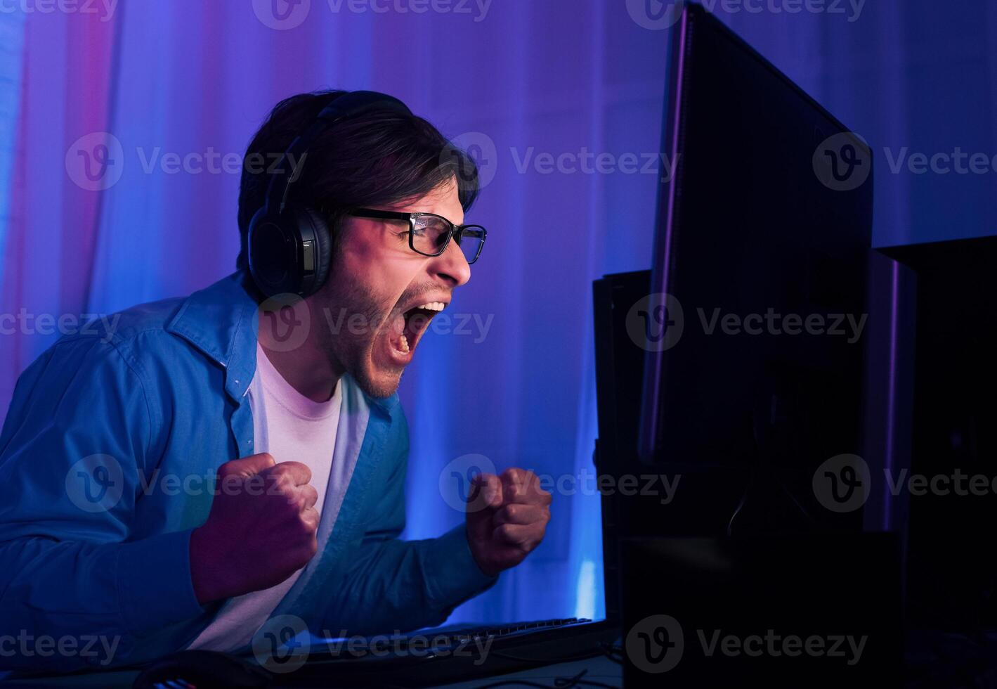 An excited gamer wearing headphones sits in front of a computer screen, cheering with his fists in the air. The gamers face is lit by blue and purple lights in the background. photo