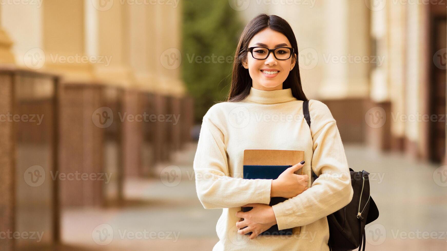 Asian young woman stands confidently on a university campus during the day, smiling while holding a couple of books close to her chest. She is wearing glasses photo