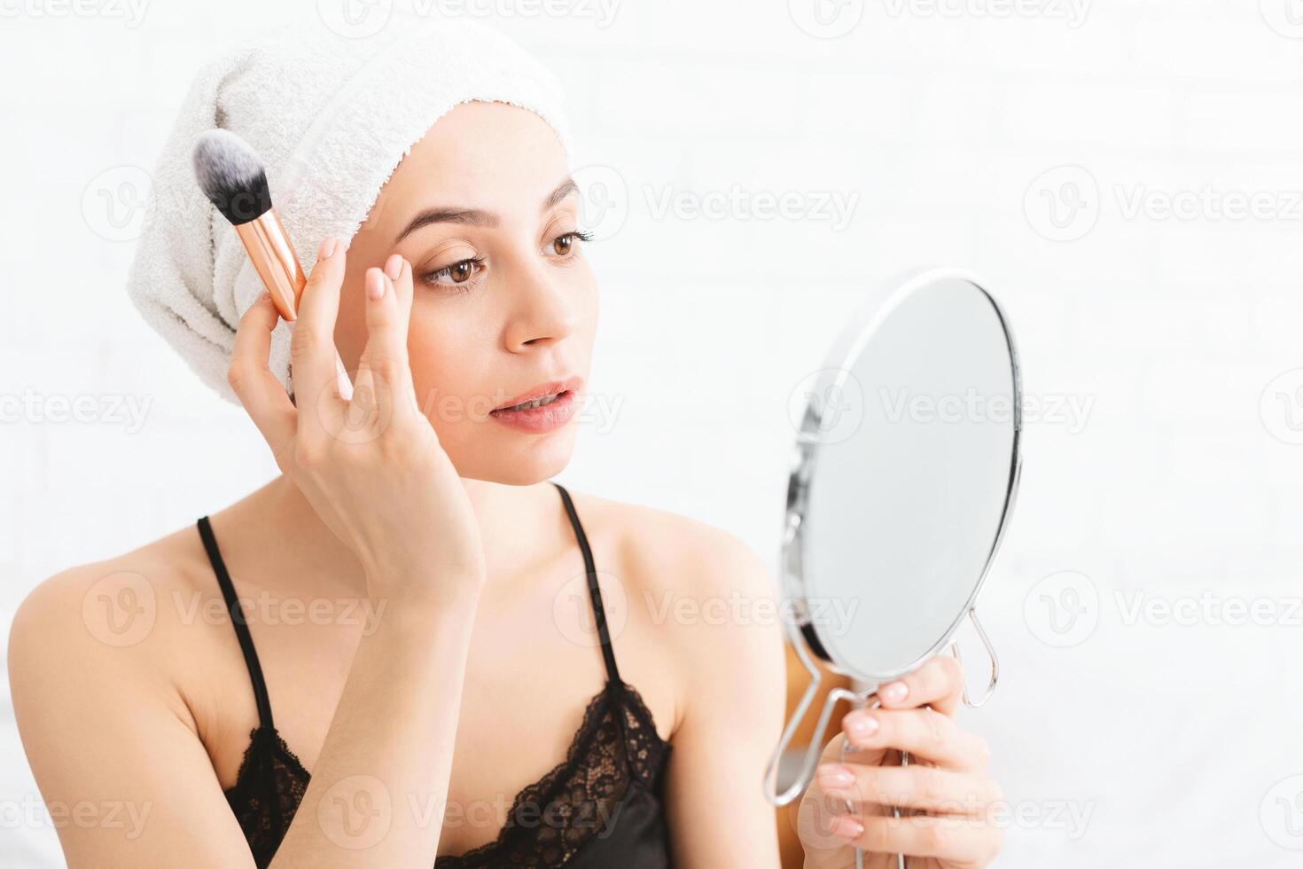 A young woman, with a towel wrapped around her hair, delicately applies makeup to her face using a brush as she intently gazes at her reflection in a handheld mirror. photo