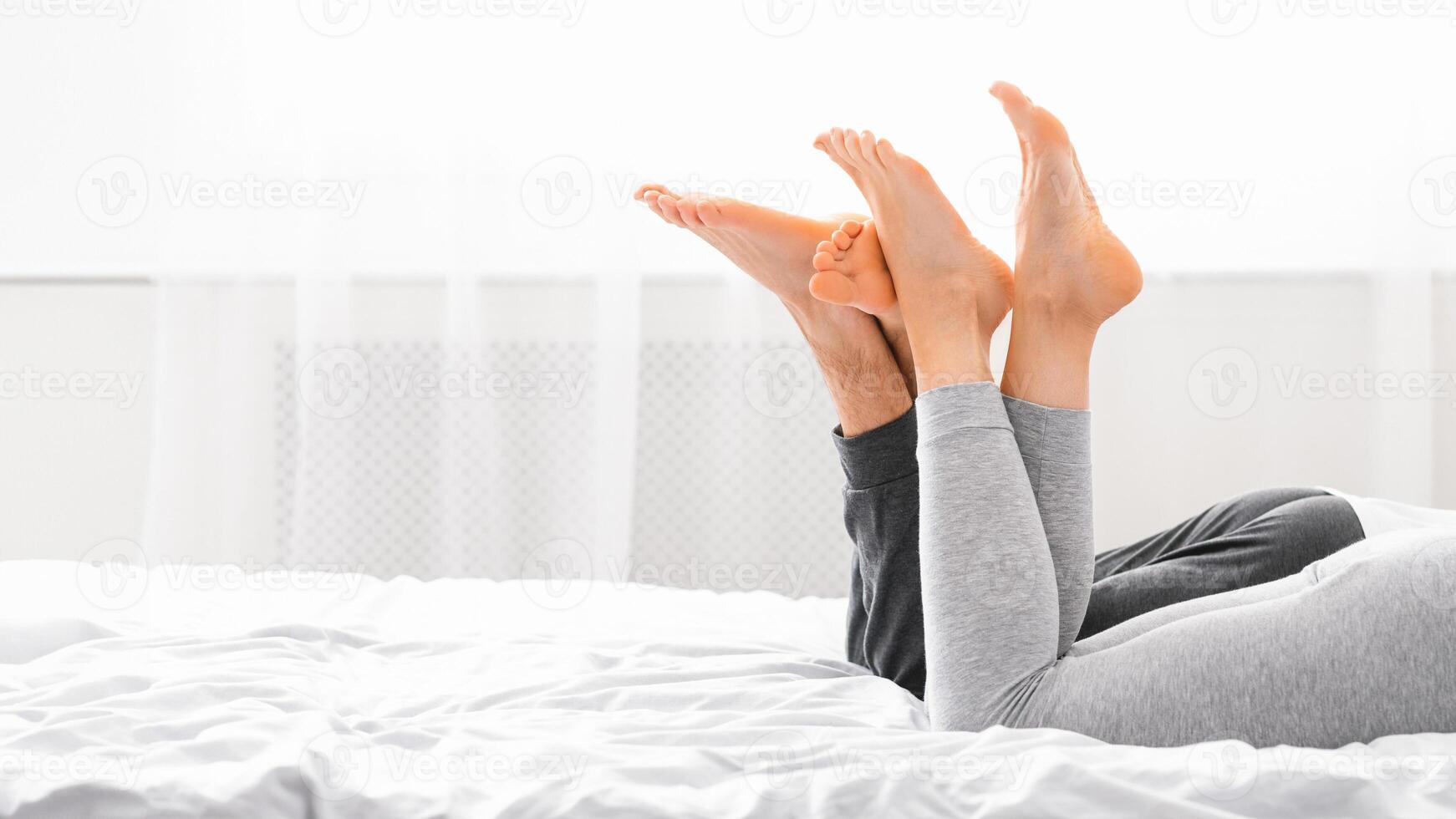 A couple lies in bed together, both with their feet raised in the air, seemingly enjoying each others company in the morning sunlight, copy space photo