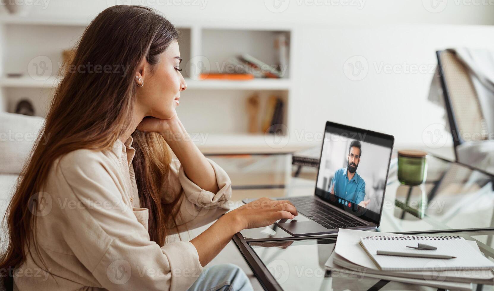 A young woman sits at a desk in her home office, attentively listening to a conference on her laptop. The room is well-lit, and the woman is dressed casually in a light-colored shirt. photo