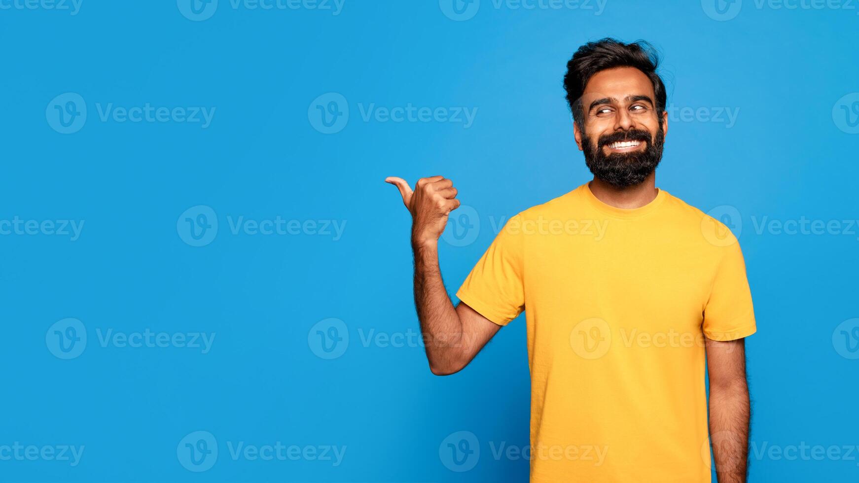 Indian man with a beard and a yellow shirt smiles and points with his thumb to copy space, standing in front of a solid blue background. photo