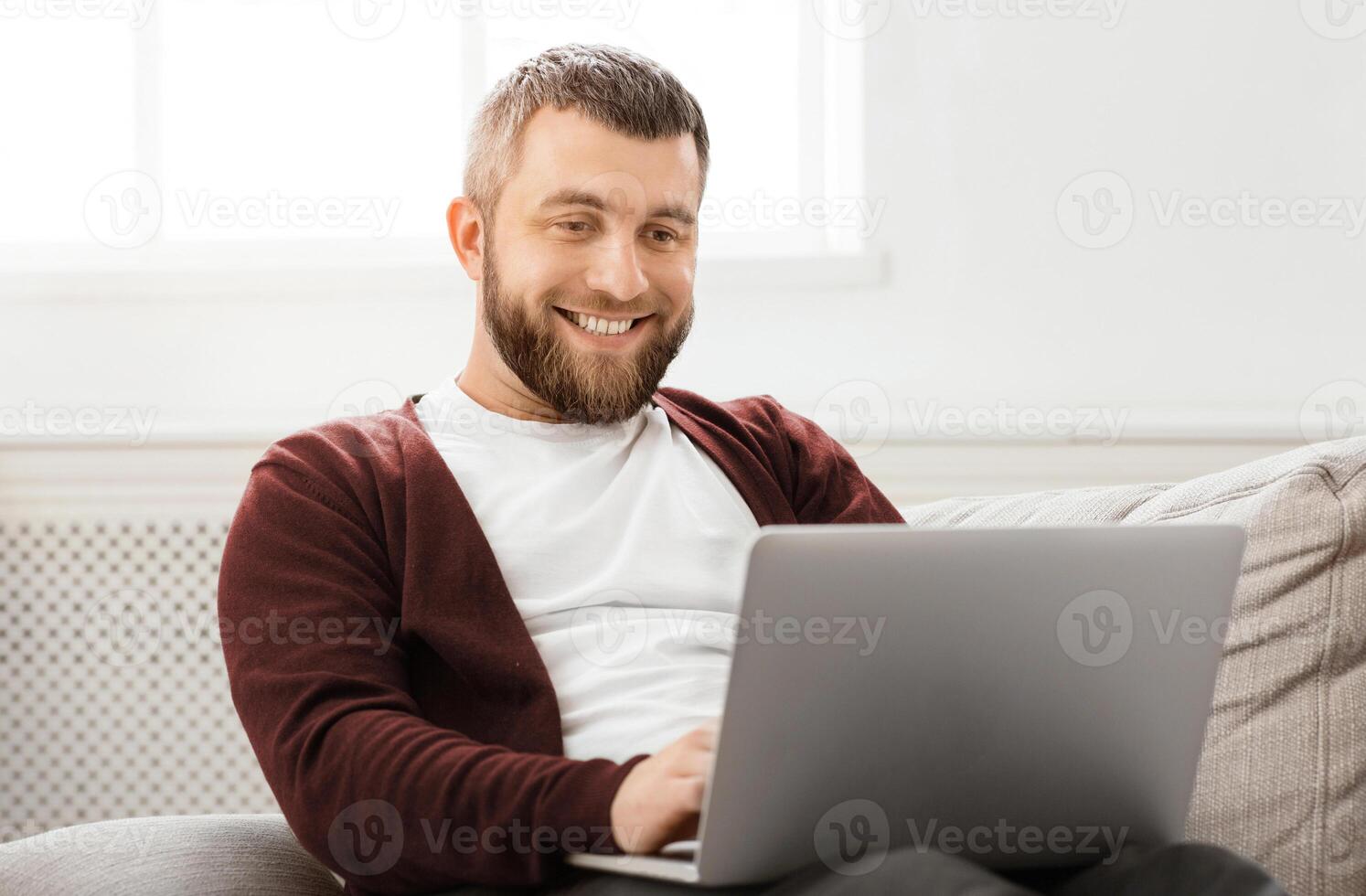 A man is seated on a comfortable couch, focused on a laptop screen in front of him. He appears engaged, possibly working or browsing. The room is well-lit, with a casual and relaxed atmosphere. photo