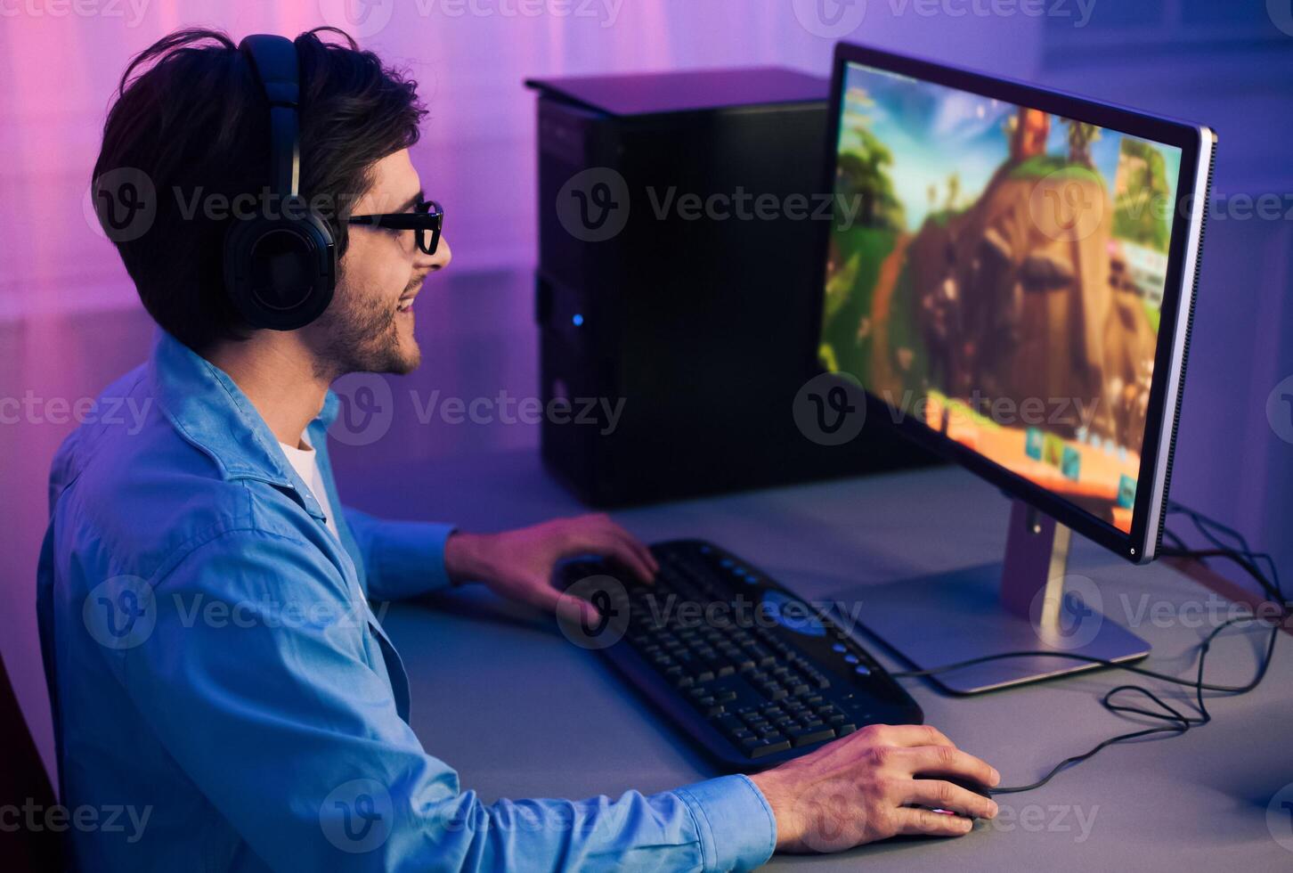 A young man sits at his computer wearing headphones and playing a game. He is smiling and seems to be enjoying himself. The game is displayed on the computer monitor and the man photo