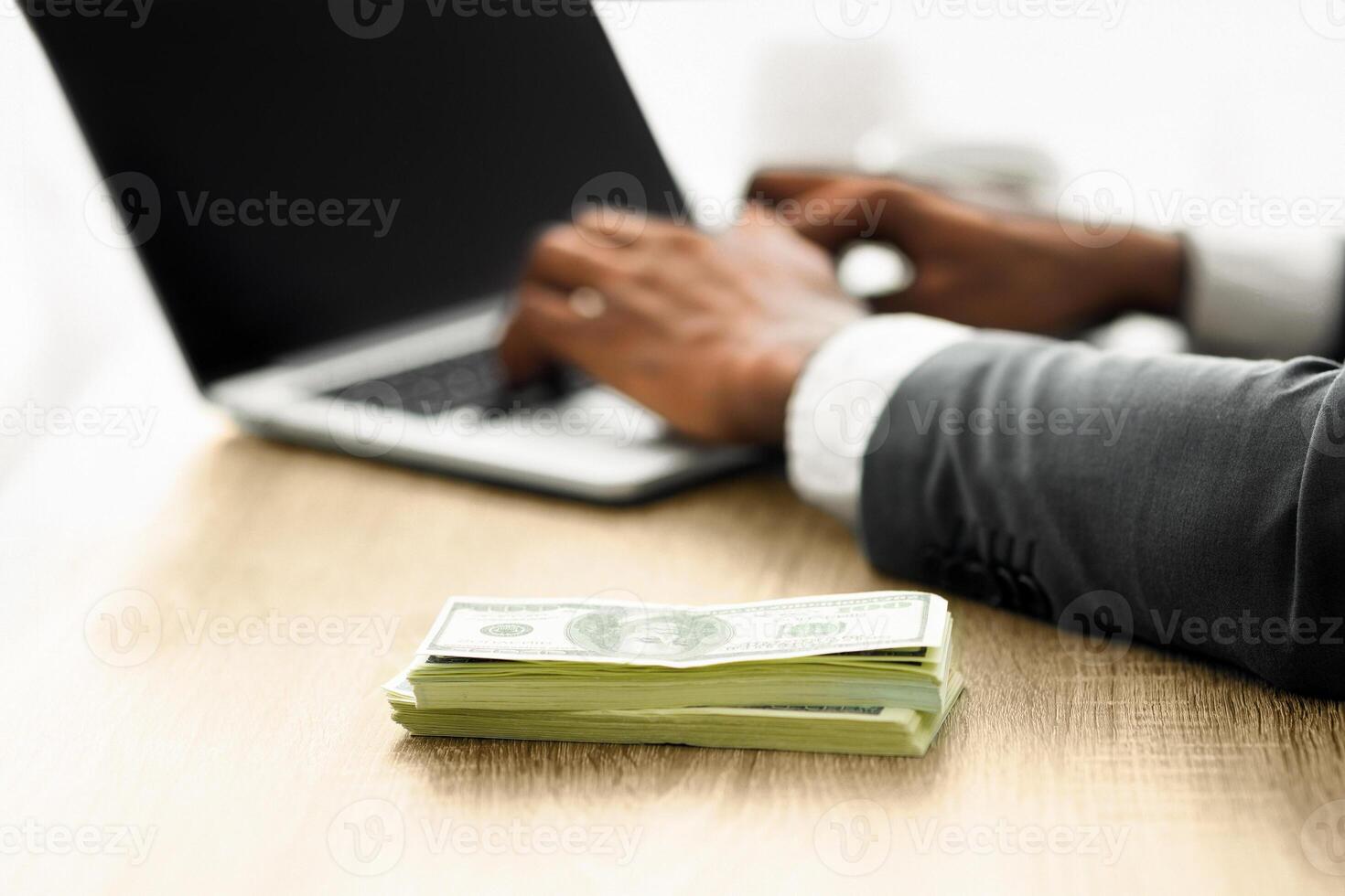 A person in a suit is working on a laptop at a desk with a stack of cash in the foreground. photo